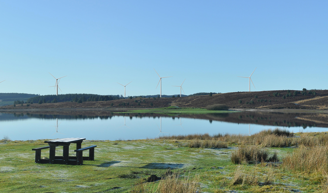 A calm lake reflecting nearby hills and wind turbines, with a wooden picnic table in the foreground under a clear blue sky.