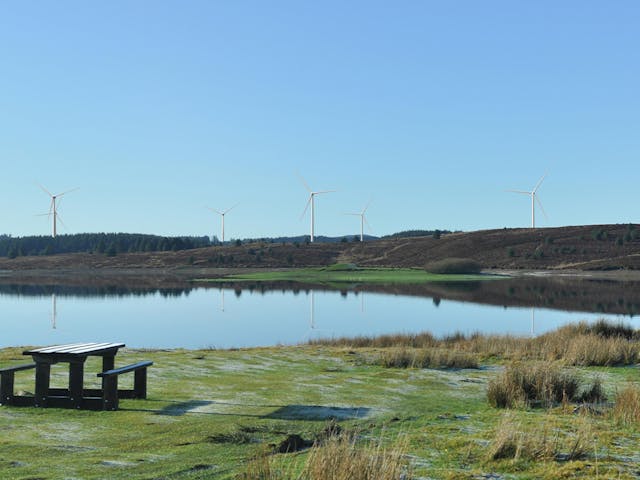 A calm lake reflecting nearby hills and wind turbines, with a wooden picnic table in the foreground under a clear blue sky.