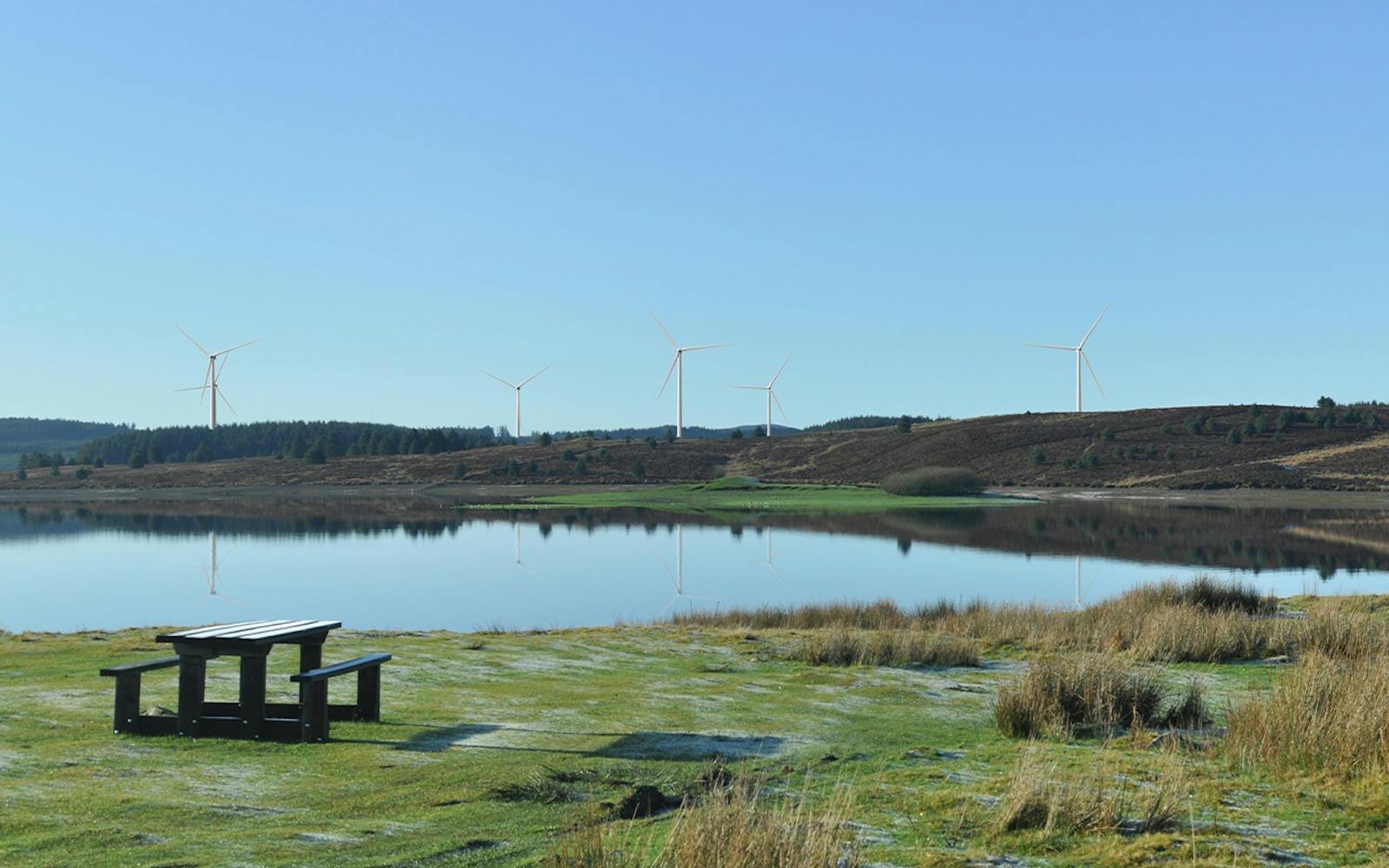 A calm lake reflecting nearby hills and wind turbines, with a wooden picnic table in the foreground under a clear blue sky.