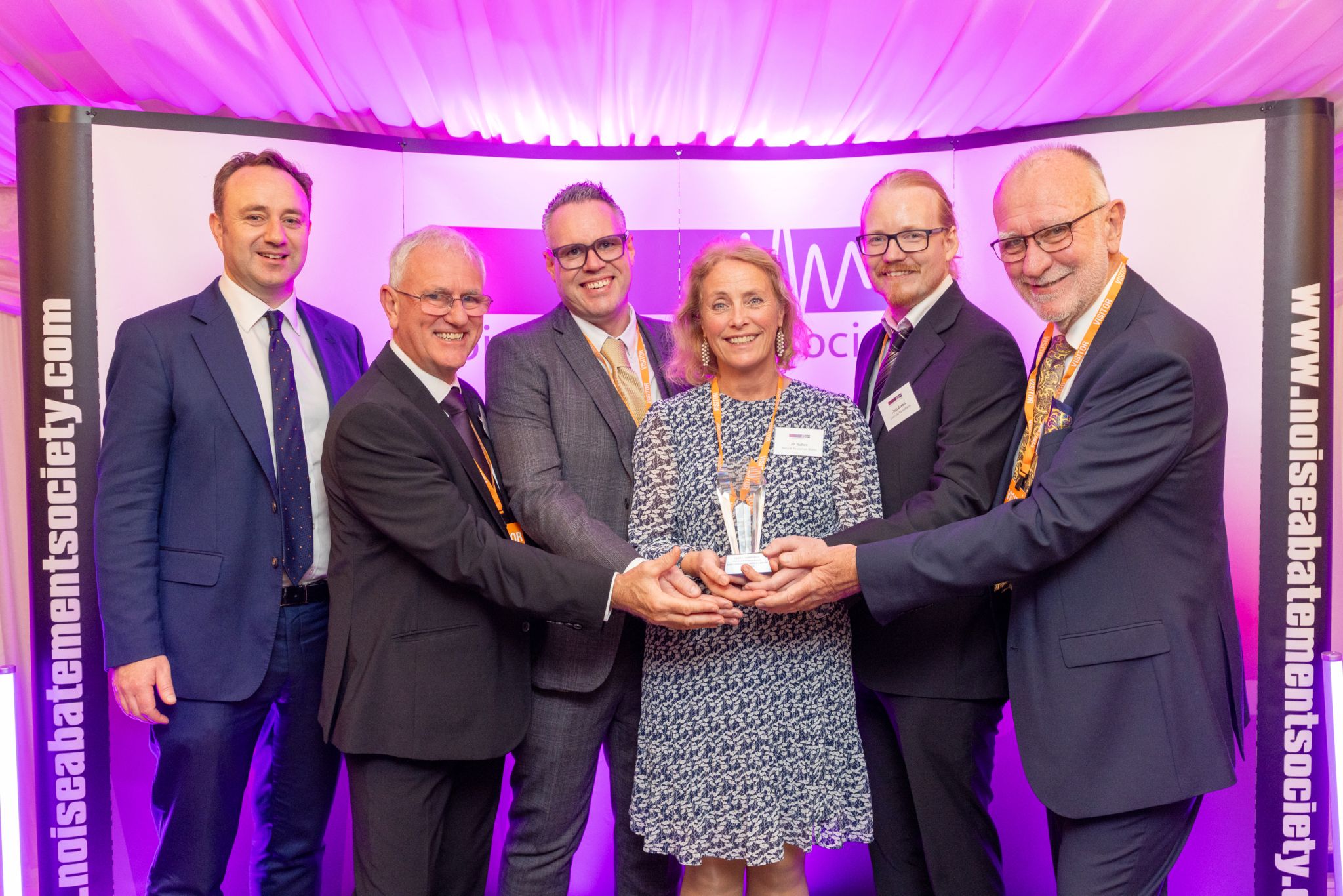 A group of six people stand together smiling and holding an award at an event hosted by the Noise Abatement Society. They are dressed in business attire and posed in front of a purple-lit backdrop displaying the society’s logo and website.