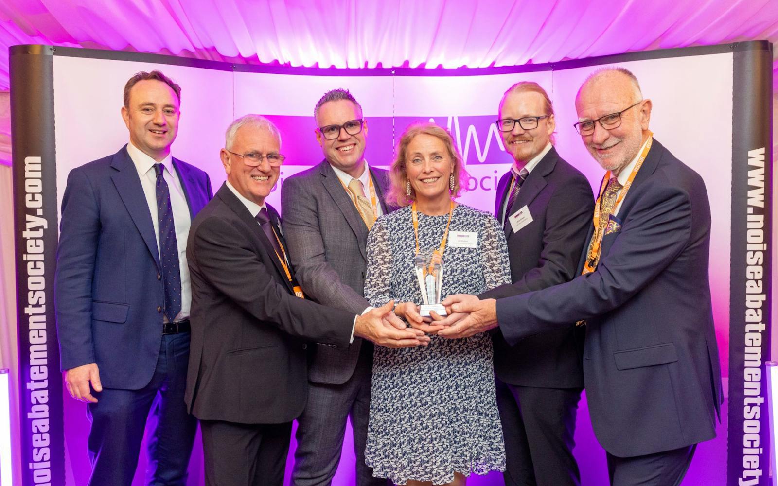 A group of six people stand together smiling and holding an award at an event hosted by the Noise Abatement Society. They are dressed in business attire and posed in front of a purple-lit backdrop displaying the society’s logo and website.