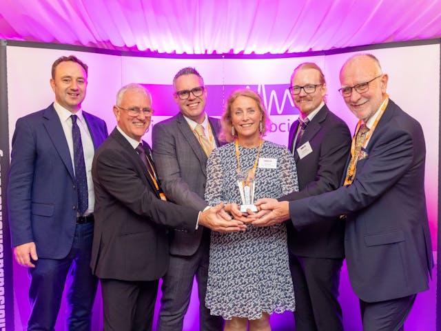 A group of six people stand together smiling and holding an award at an event hosted by the Noise Abatement Society. They are dressed in business attire and posed in front of a purple-lit backdrop displaying the society’s logo and website.