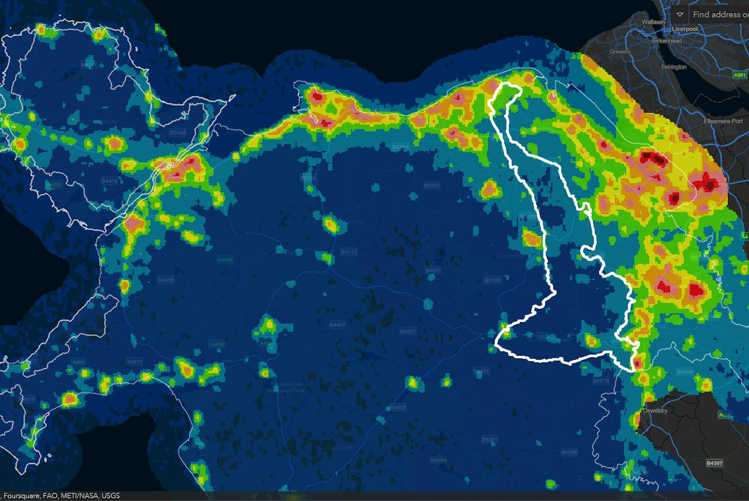 A heatmap of Wales and surrounding areas showing light pollution levels. Brighter colours indicate higher light pollution, with red and yellow concentrated along the north coast and near major towns. Dark blue areas represent darker skies, mainly across mid and west Wales. White boundary lines highlight regional borders.