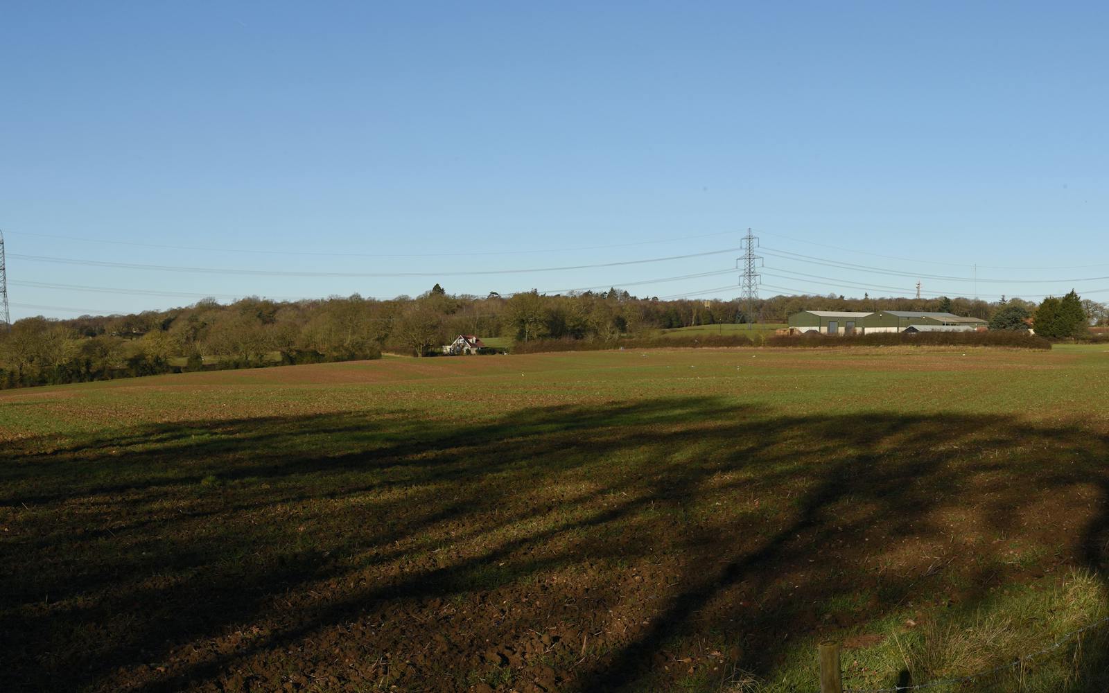 Rural landscape with a large open field under a clear blue sky, featuring patches of green vegetation, distant trees, a barn-like structure, and tall electrical transmission towers with power lines stretching across the scene.