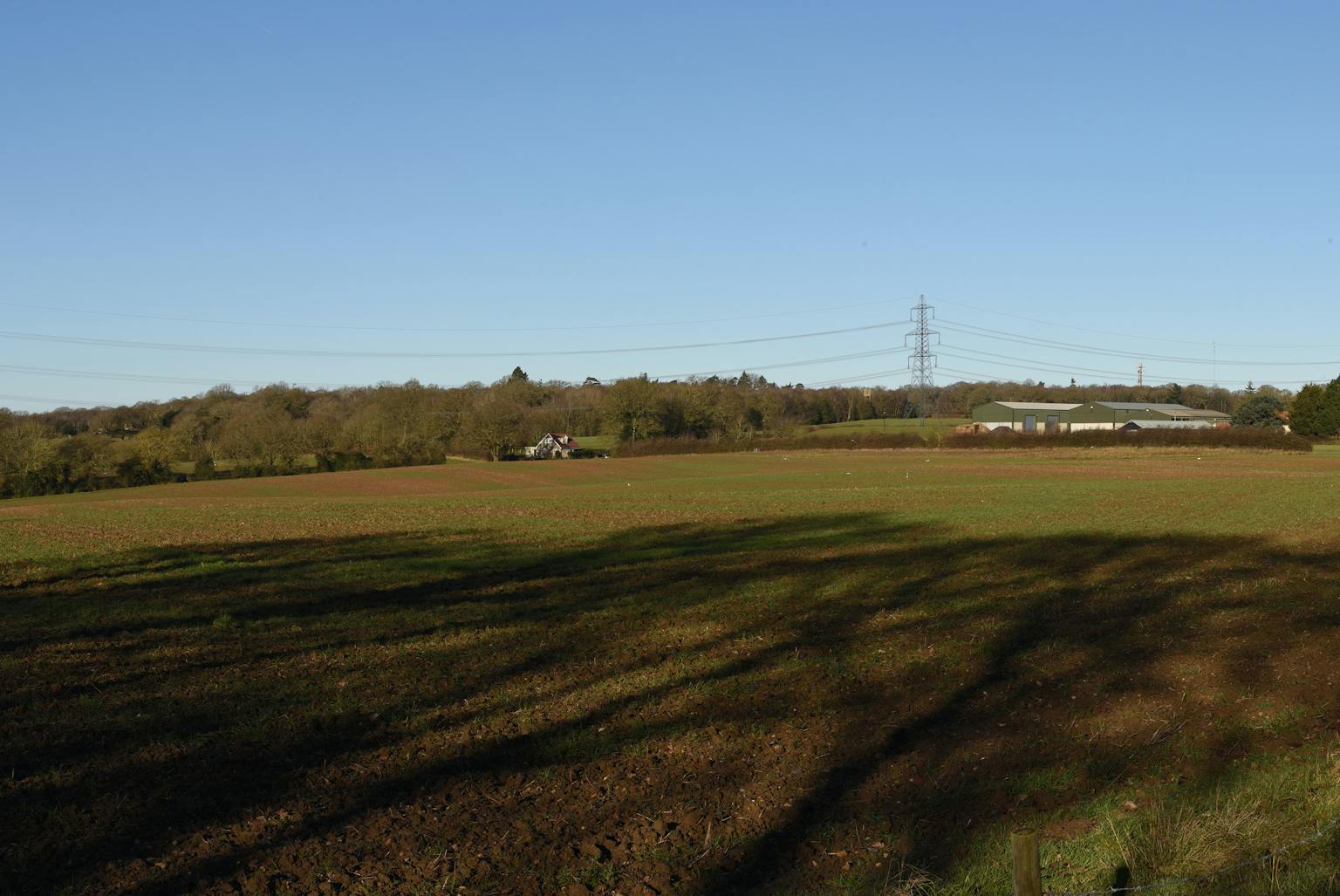 Rural landscape with a large open field under a clear blue sky, featuring patches of green vegetation, distant trees, a barn-like structure, and tall electrical transmission towers with power lines stretching across the scene.