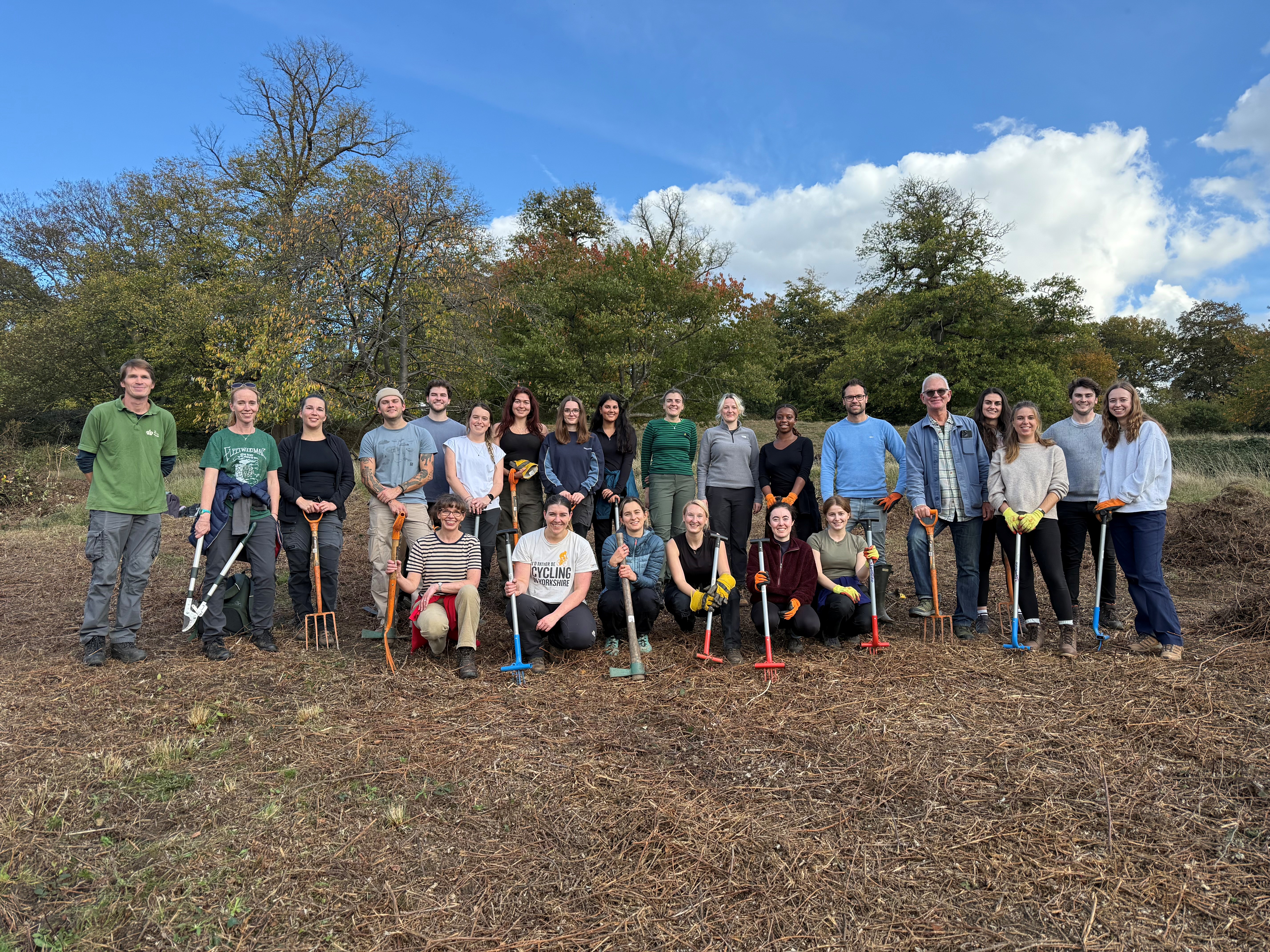 A large group of LUC volunteers pose together in Richmond Park, smiling and holding gardening tools after a successful habitat restoration day under a bright autumn sky.