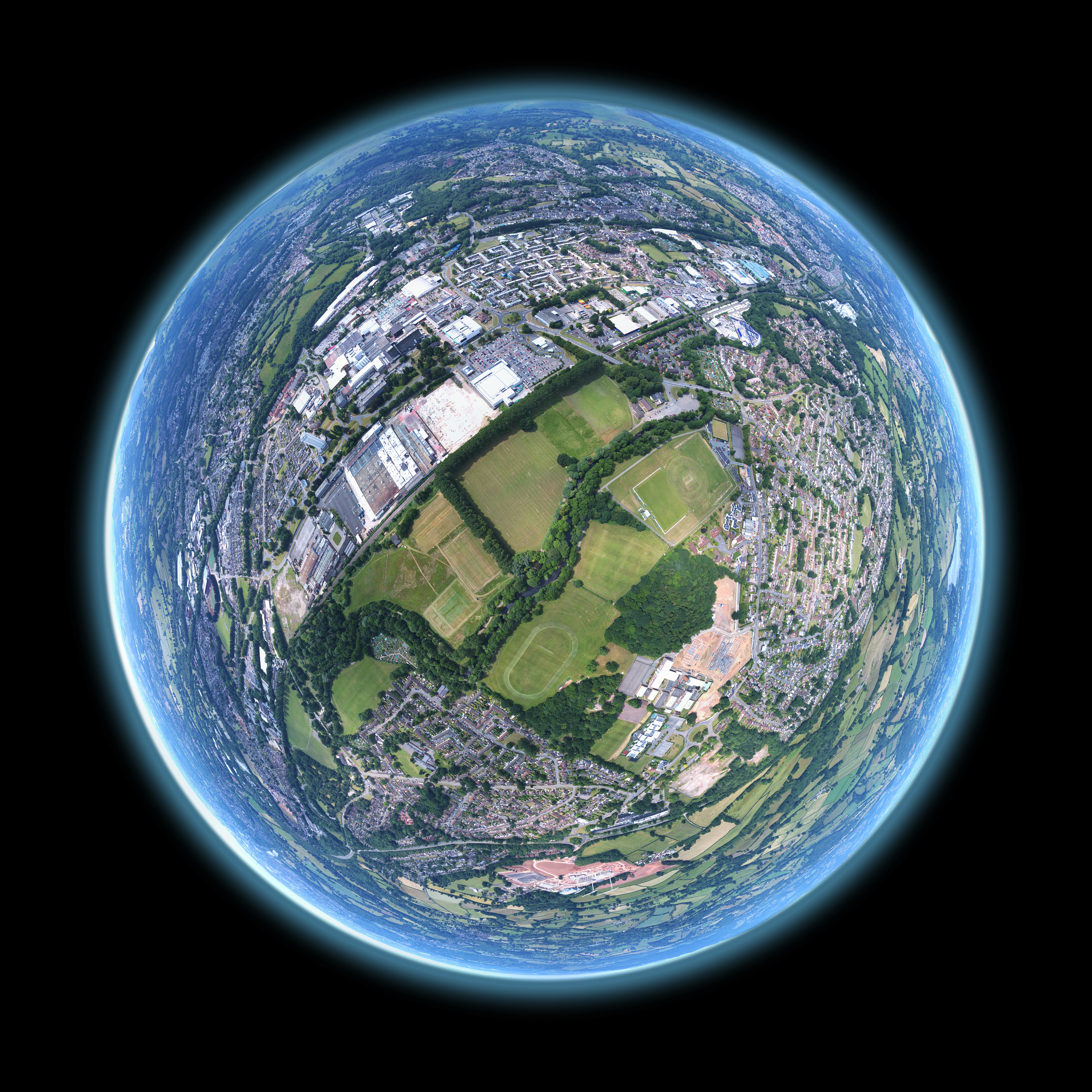 A circular aerial photo, often called a 'tiny planet' or stereographic projection, showing a mix of urban development, large commercial buildings and parking lots, residential areas, and green spaces, including several sports fields, surrounded by rolling hills and countryside. The entire image is contained within a glowing blue-edged circle against a black background.