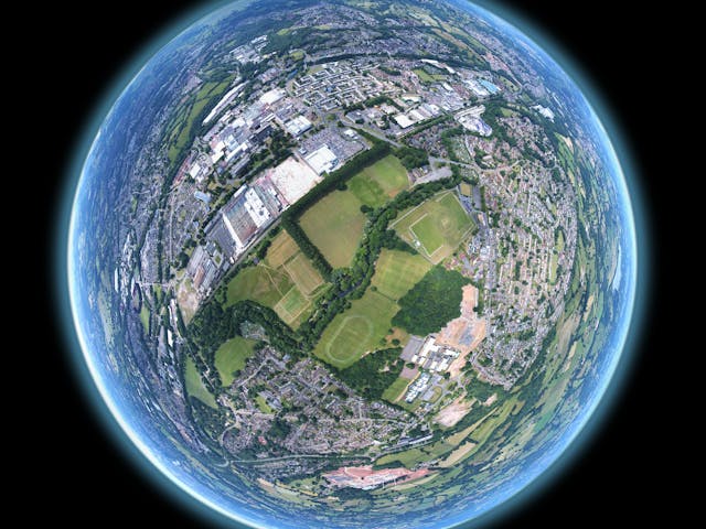 A circular aerial photo, often called a 'tiny planet' or stereographic projection, showing a mix of urban development, large commercial buildings and parking lots, residential areas, and green spaces, including several sports fields, surrounded by rolling hills and countryside. The entire image is contained within a glowing blue-edged circle against a black background.