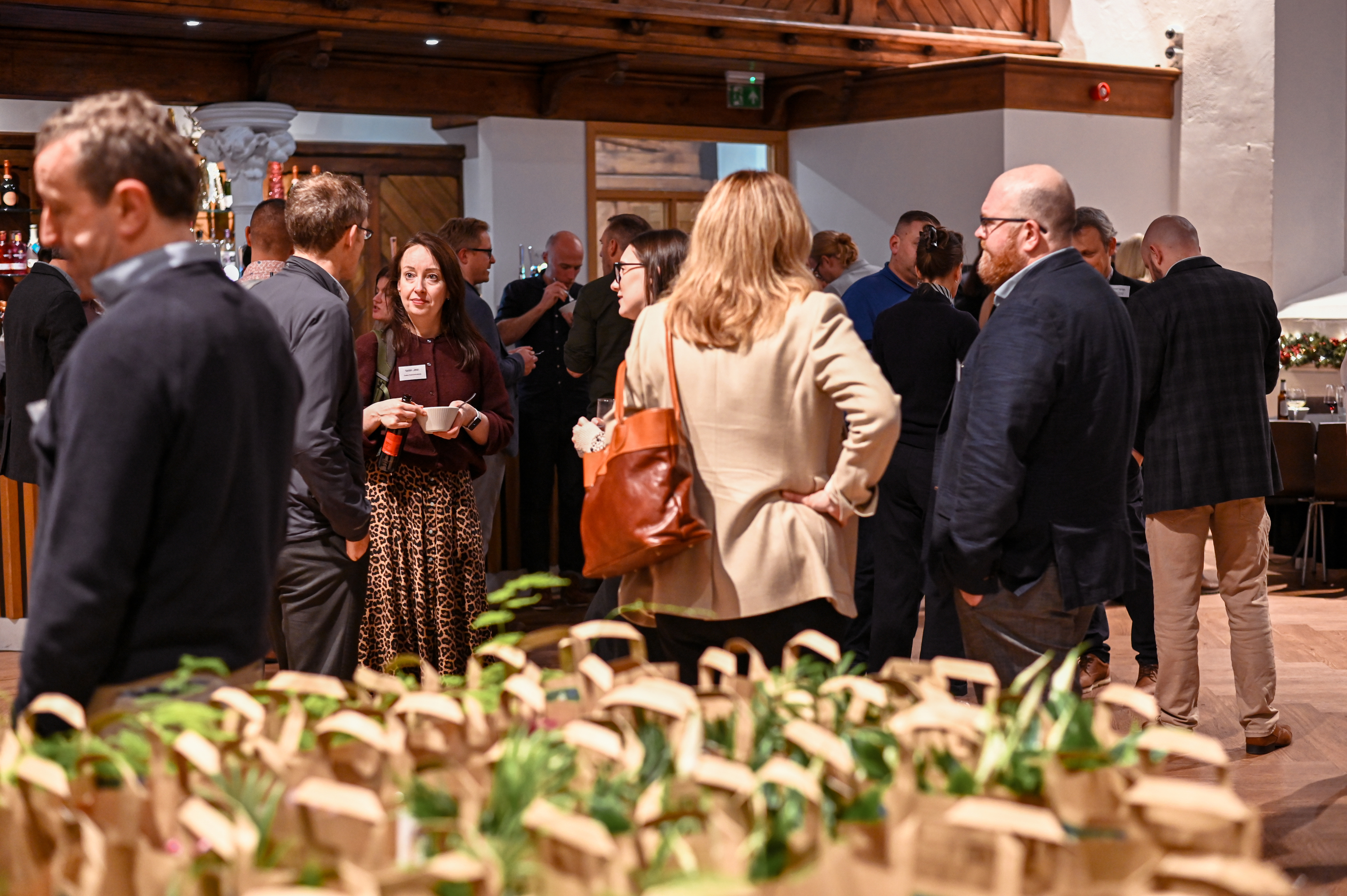 A professional networking event taking place indoors. Guests are standing in small groups, chatting, with many small potted plants displayed on a table in the blurred foreground.