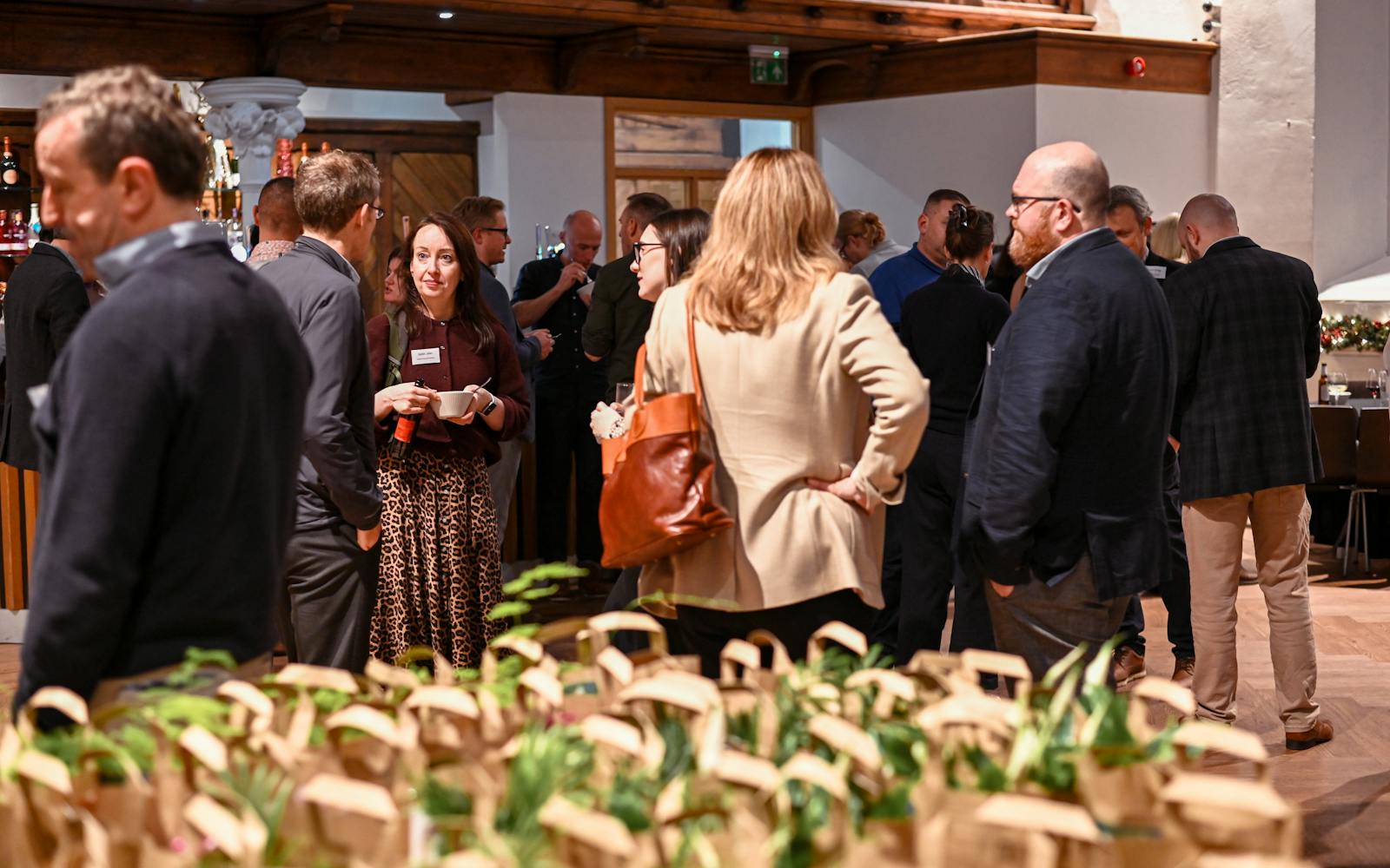 A professional networking event taking place indoors. Guests are standing in small groups, chatting, with many small potted plants displayed on a table in the blurred foreground.