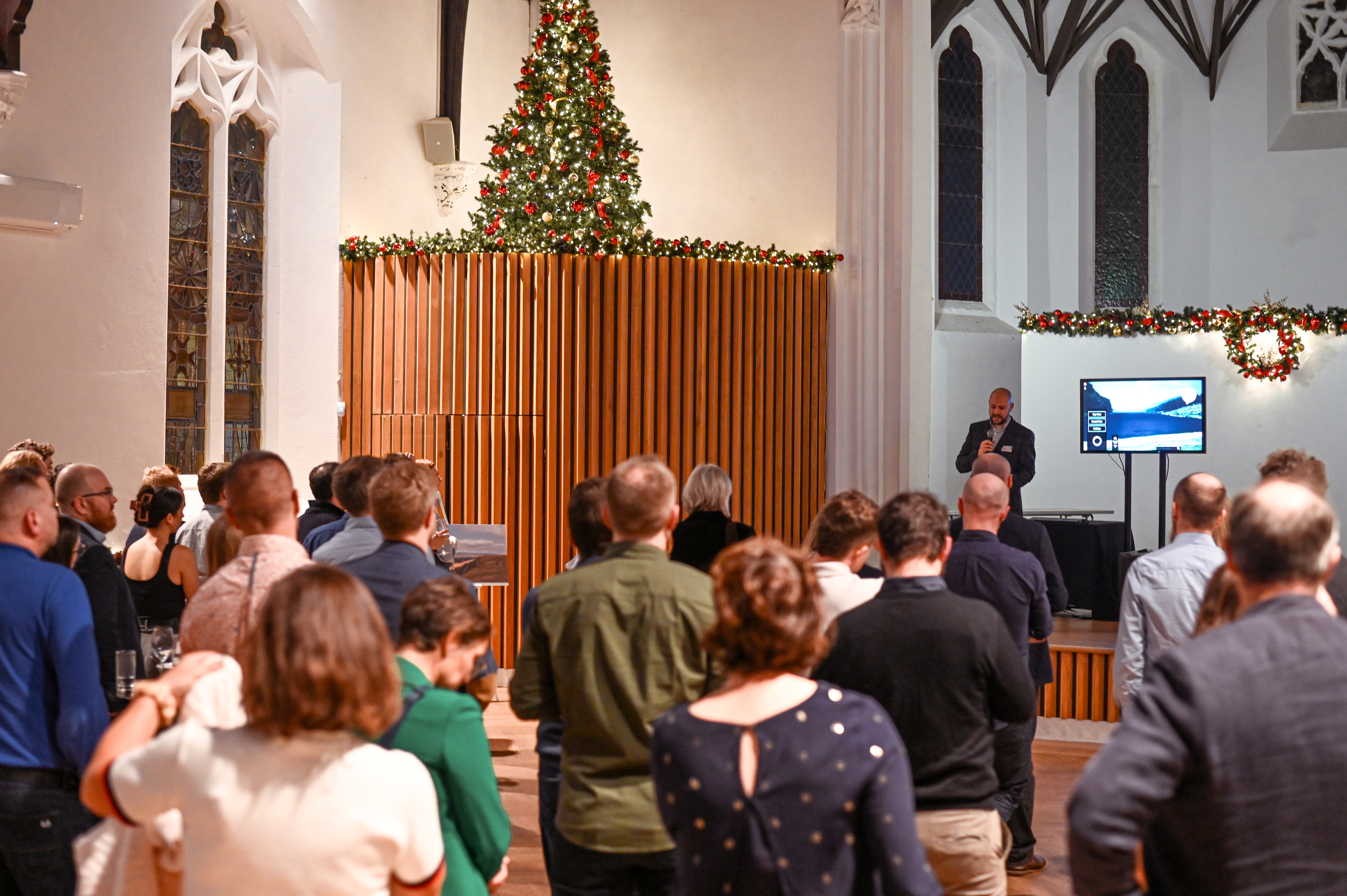 A crowd of people faces forward, listening to a man speaking into a microphone near a large screen displaying a presentation slide. The event is taking place indoors in a hall or church with tall windows, holiday garlands, and a Christmas tree visible above a wooden wall.