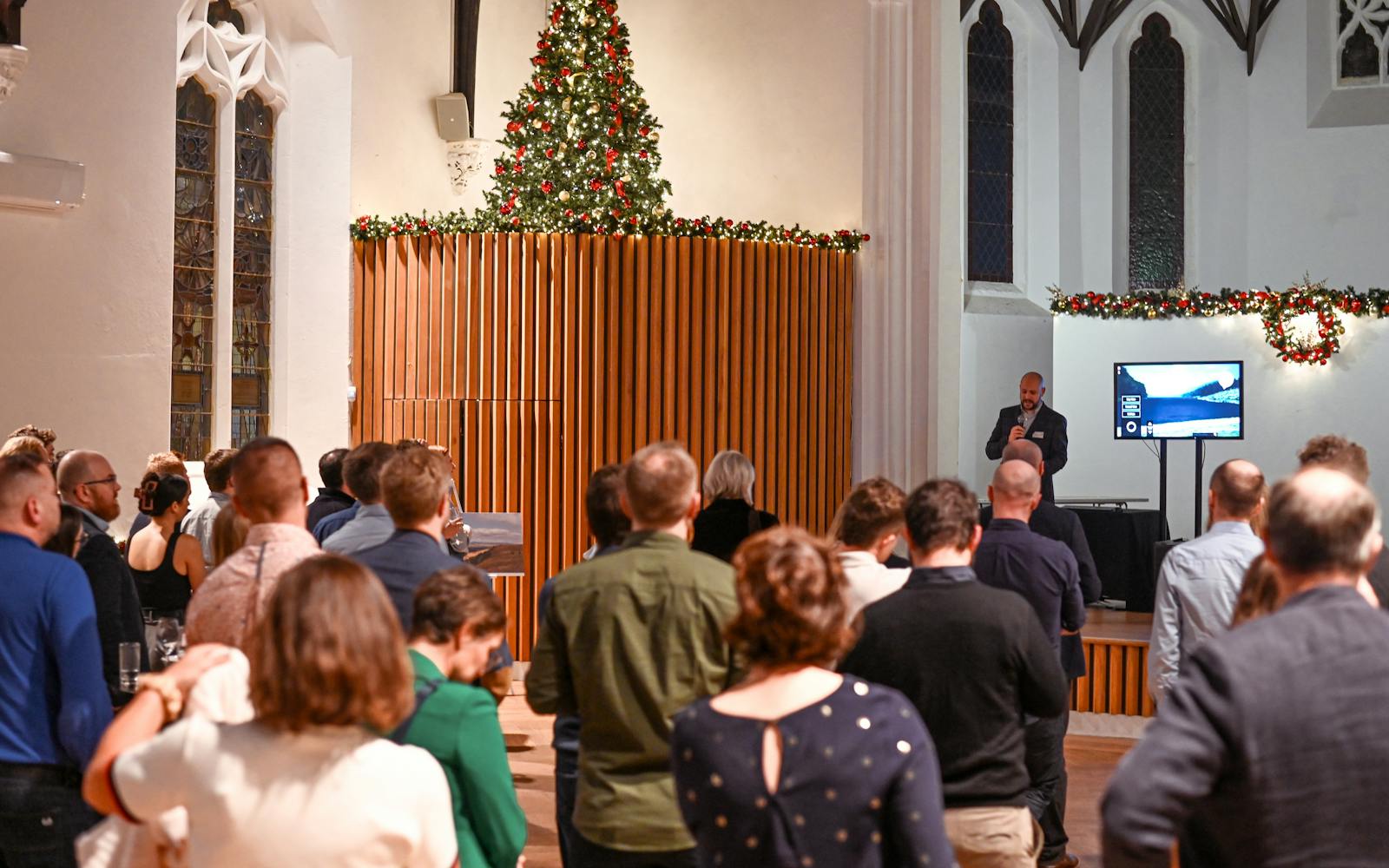 A crowd of people faces forward, listening to a man speaking into a microphone near a large screen displaying a presentation slide. The event is taking place indoors in a hall or church with tall windows, holiday garlands, and a Christmas tree visible above a wooden wall.