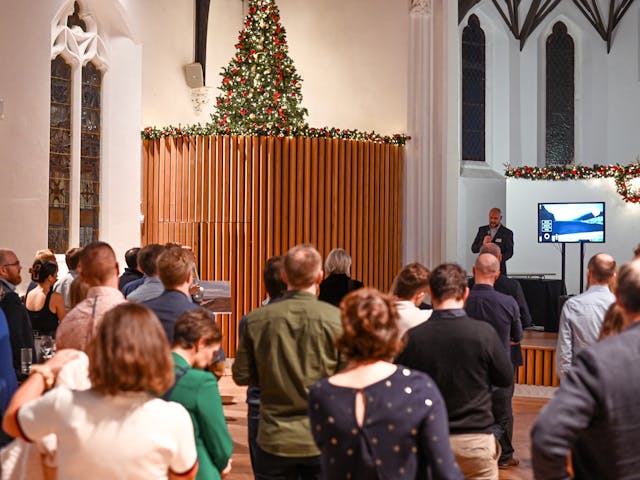 A crowd of people faces forward, listening to a man speaking into a microphone near a large screen displaying a presentation slide. The event is taking place indoors in a hall or church with tall windows, holiday garlands, and a Christmas tree visible above a wooden wall.