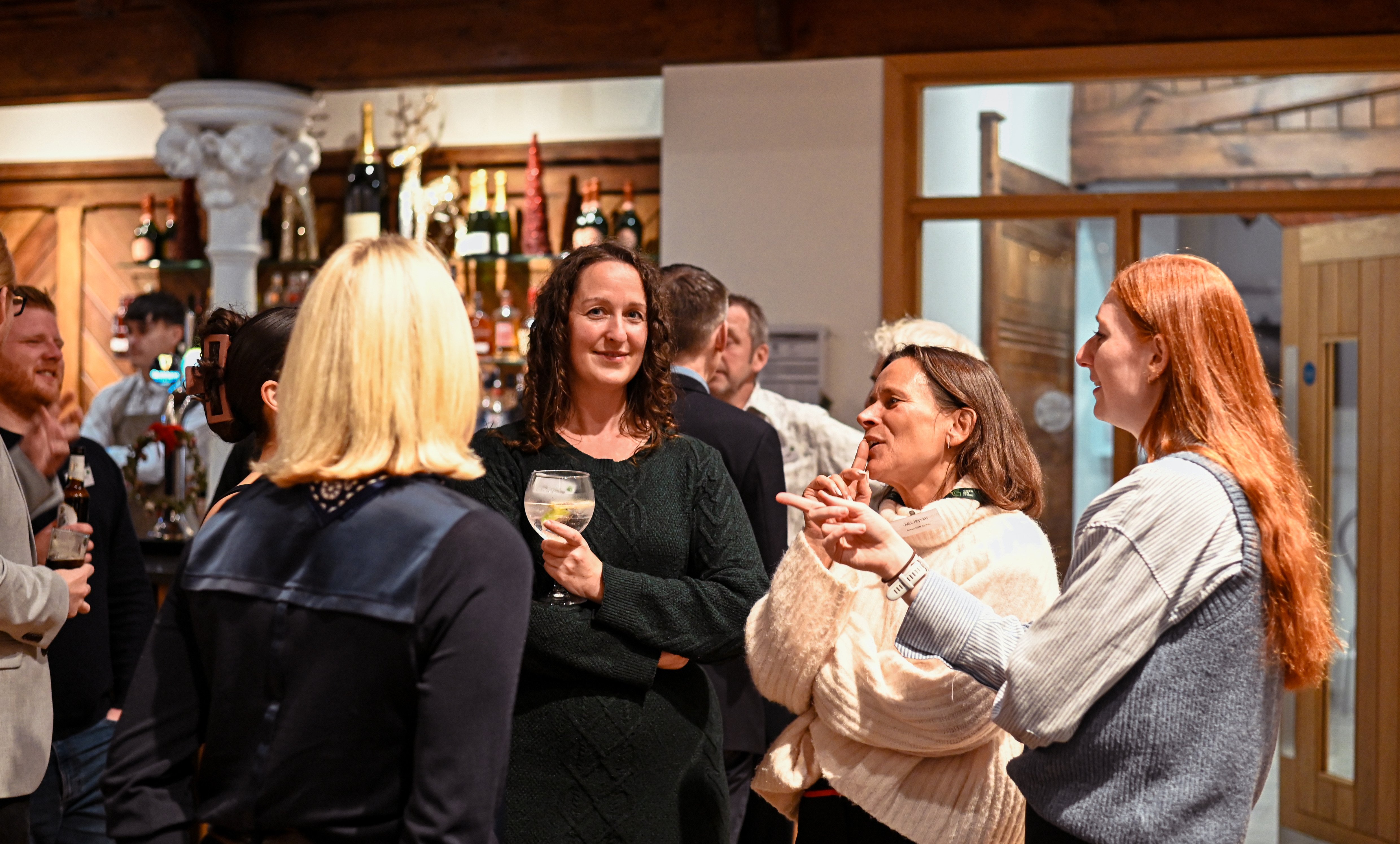A group of attendees, primarily women, stand in a close circle talking and laughing during an indoor networking event. One woman in a dark green sweater holds a cocktail, while another woman in a cream-colored chunky cardigan actively gestures with her hands as she speaks. A bar with bottles of wine and spirits is visible in the background.