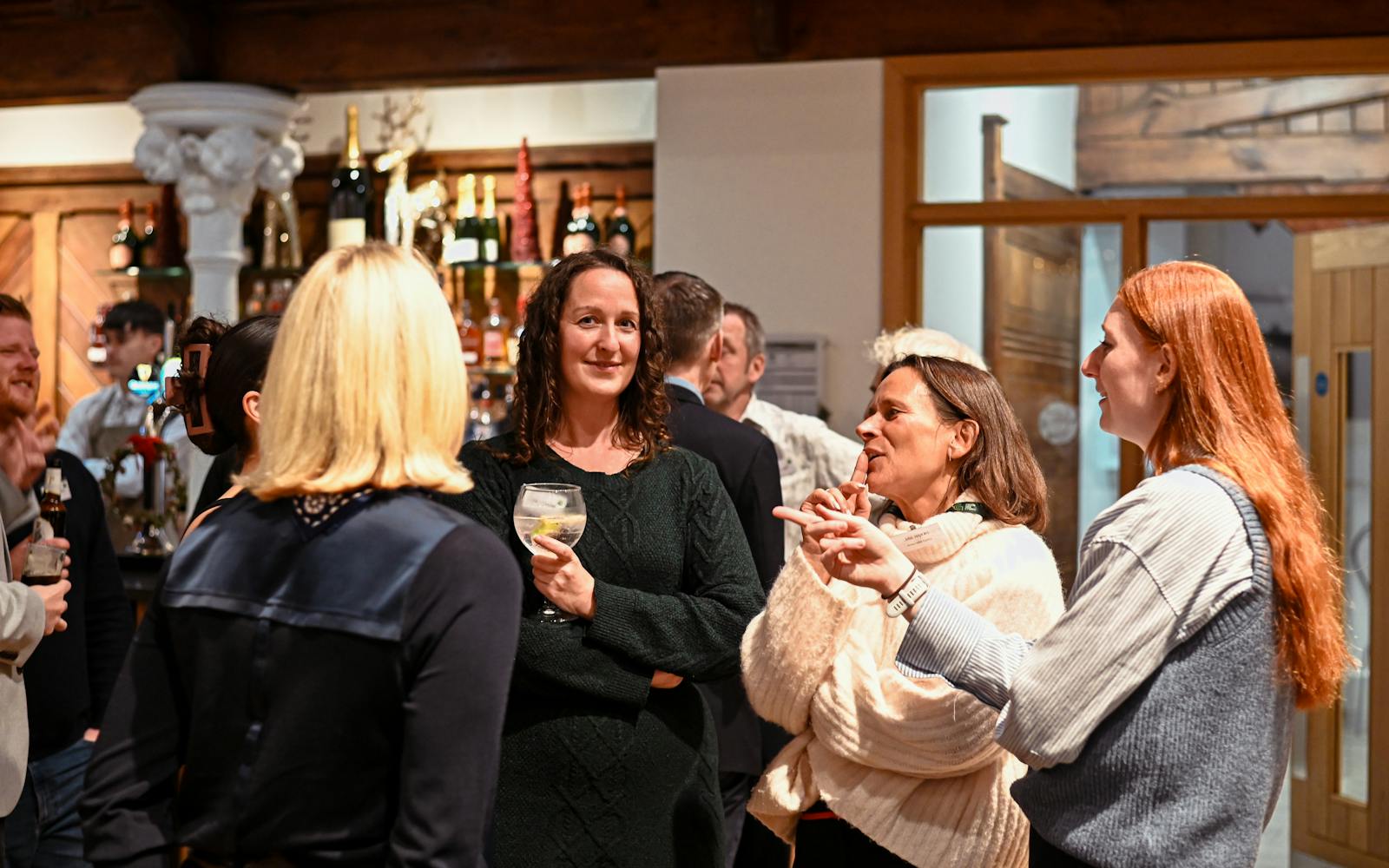 A group of attendees, primarily women, stand in a close circle talking and laughing during an indoor networking event. One woman in a dark green sweater holds a cocktail, while another woman in a cream-colored chunky cardigan actively gestures with her hands as she speaks. A bar with bottles of wine and spirits is visible in the background.