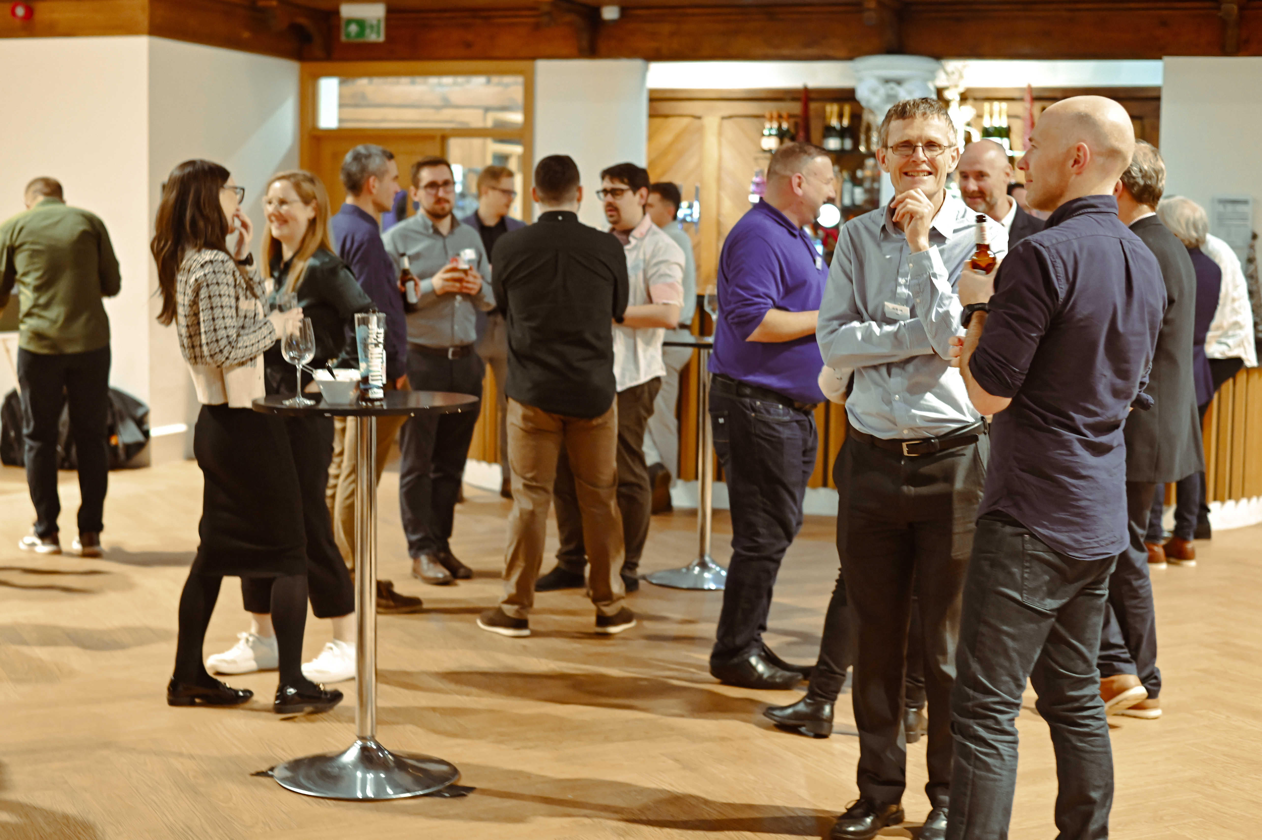 An indoor professional event where a large group of people are socializing. Two men stand in the foreground talking and smiling, one holding a bottle of beer. Other small groups are visible throughout the room, which has a wood floor and a bar area with bottles visible in the background.
