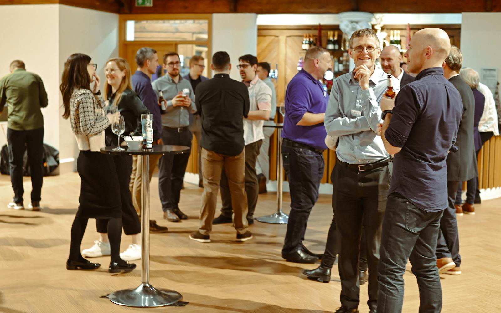 An indoor professional event where a large group of people are socializing. Two men stand in the foreground talking and smiling, one holding a bottle of beer. Other small groups are visible throughout the room, which has a wood floor and a bar area with bottles visible in the background.