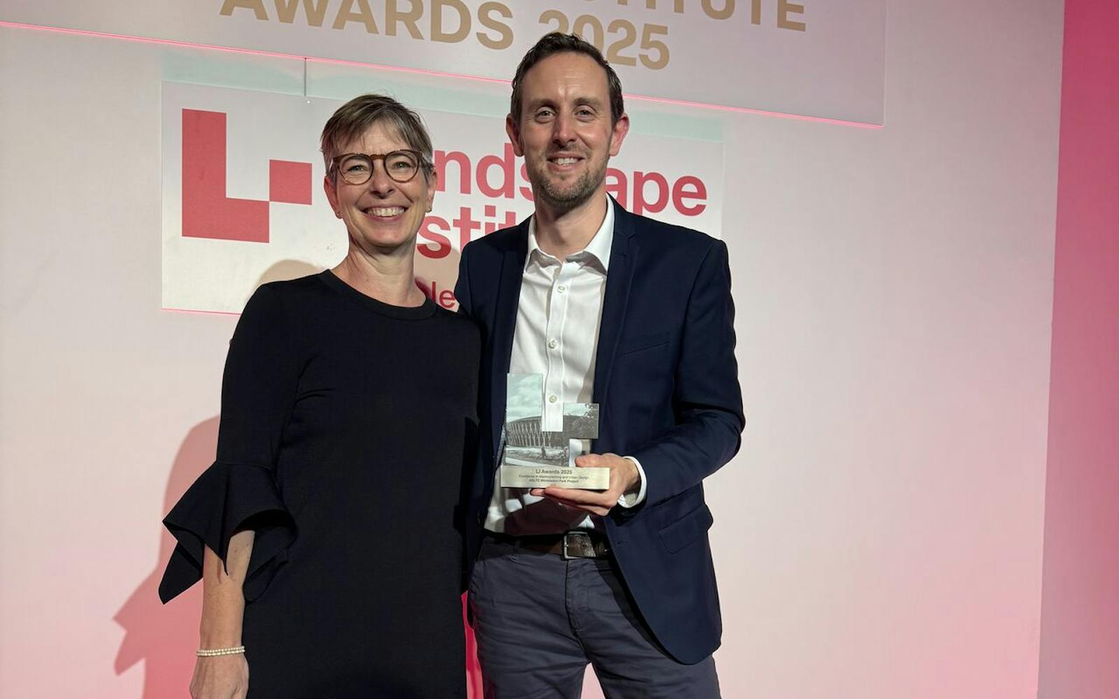 A smiling woman in a black dress, black tights, and tall black boots stands next to a man in a navy suit, white shirt, and white trainers. The man is holding a clear, geometric award trophy. They are on a stage with a bright pink backdrop. Above them is a sign that reads, "LANDSCAPE INSTITUTE AWARDS 2025," with the Landscape Institute logo below.