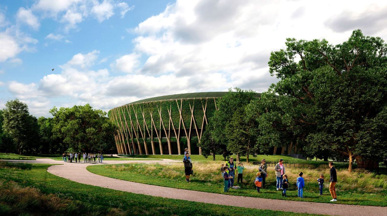 A rendering of a large, circular building surrounded by a park-like setting. The building's exterior is covered in a green wall of vegetation, supported by a striking pattern of light brown, diagonal wooden structural elements that resemble large intersecting branches. A curved, paved pathway cuts through the foreground, which is lined with lush grass and mature trees on both sides. Several small groups of adults and children are visible along the path and in the grassy area, suggesting a public space. The sky is partly cloudy and blue.