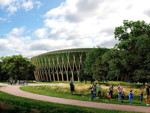 A rendering of a large, circular building surrounded by a park-like setting. The building's exterior is covered in a green wall of vegetation, supported by a striking pattern of light brown, diagonal wooden structural elements that resemble large intersecting branches. A curved, paved pathway cuts through the foreground, which is lined with lush grass and mature trees on both sides. Several small groups of adults and children are visible along the path and in the grassy area, suggesting a public space. The sky is partly cloudy and blue.