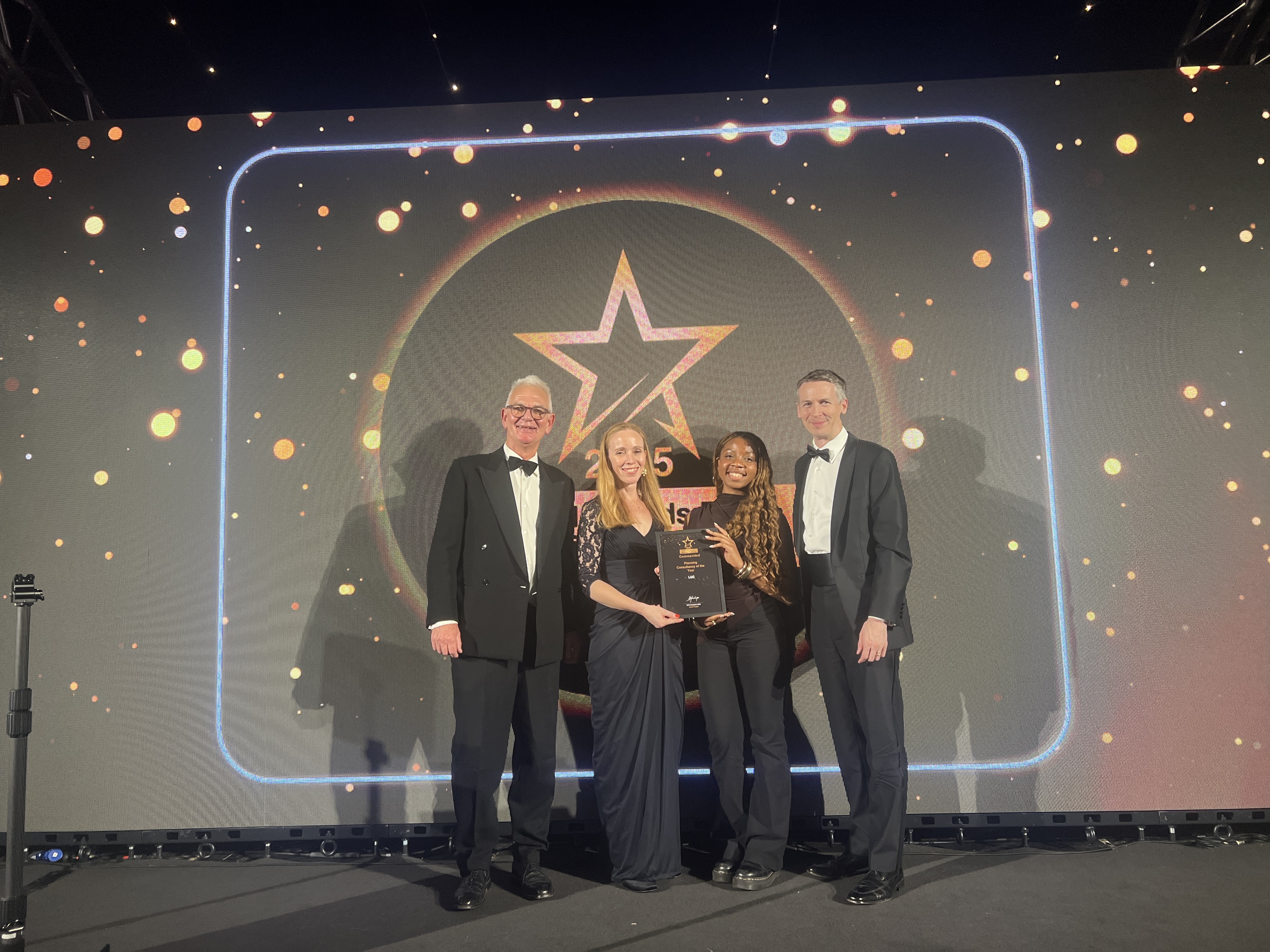 A photograph of four people on a stage, all dressed in formal attire, standing in front of a large screen displaying a star logo and bright lights. They are holding a framed award.