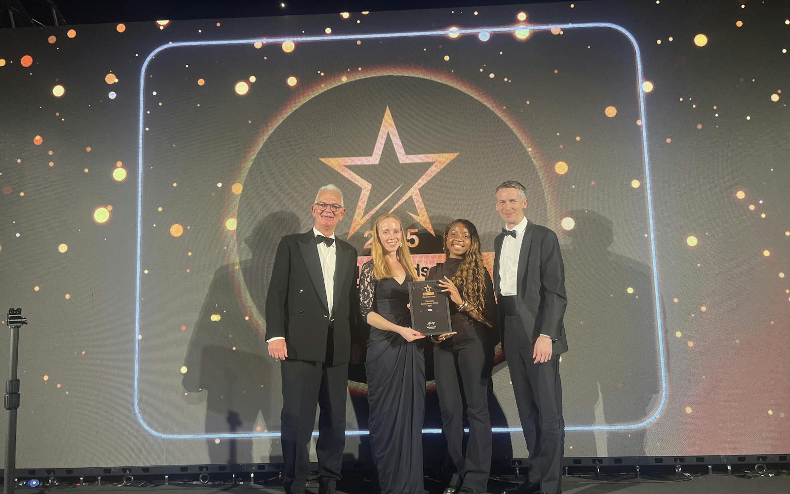 A photograph of four people on a stage, all dressed in formal attire, standing in front of a large screen displaying a star logo and bright lights. They are holding a framed award.