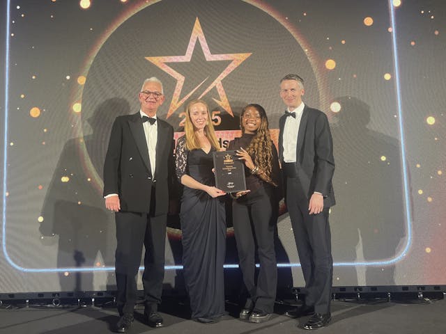 A photograph of four people on a stage, all dressed in formal attire, standing in front of a large screen displaying a star logo and bright lights. They are holding a framed award.