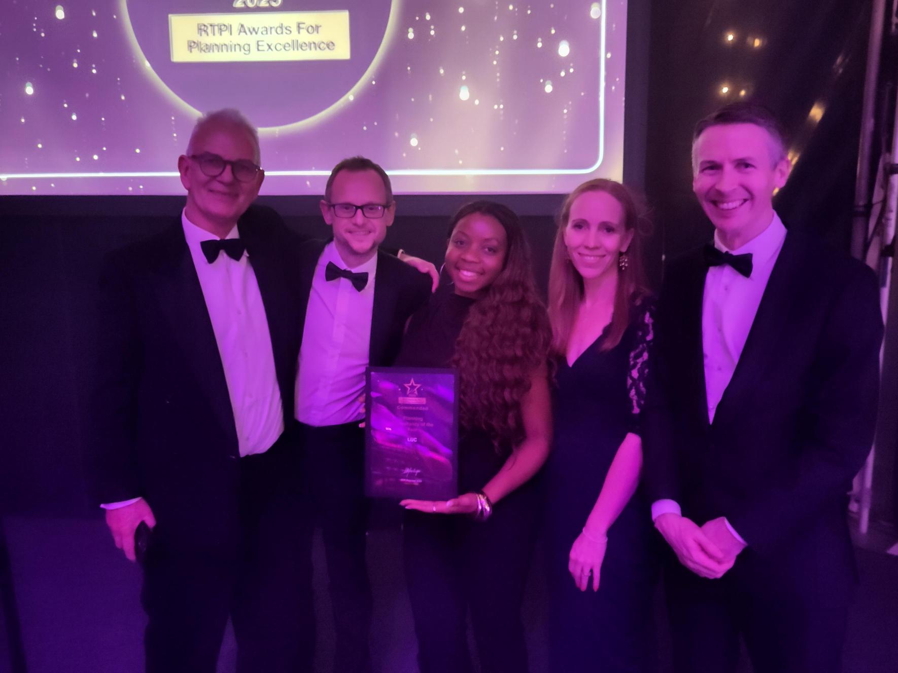 A group photograph of five people, all dressed in formal attire (tuxedos/dark gowns), standing side-by-side on a stage or platform. The person in the centre is holding a framed award. Behind them is a large screen displaying the RTPI Awards For Planning Excellence logo and text.