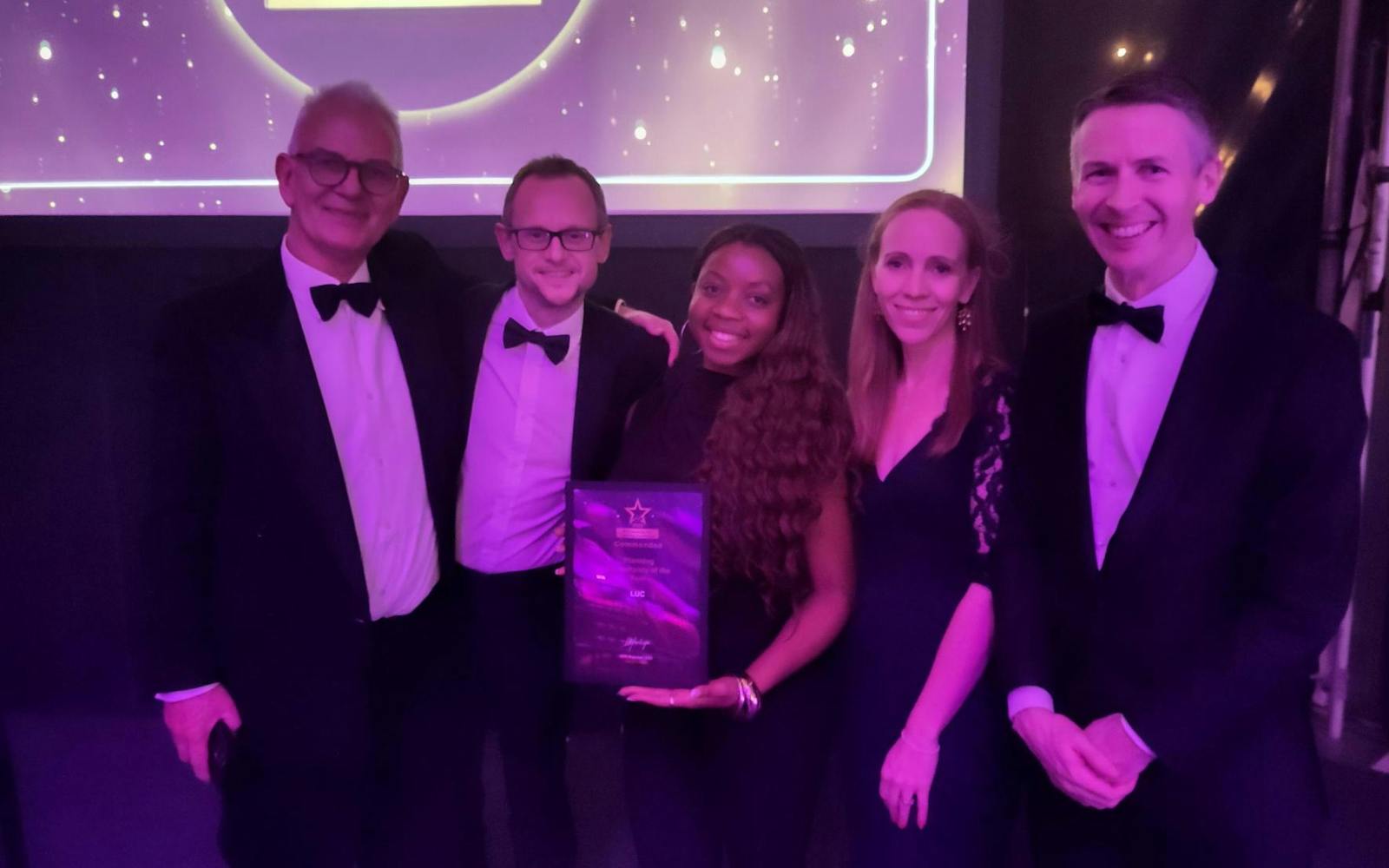 A group photograph of five people, all dressed in formal attire (tuxedos/dark gowns), standing side-by-side on a stage or platform. The person in the centre is holding a framed award. Behind them is a large screen displaying the RTPI Awards For Planning Excellence logo and text.
