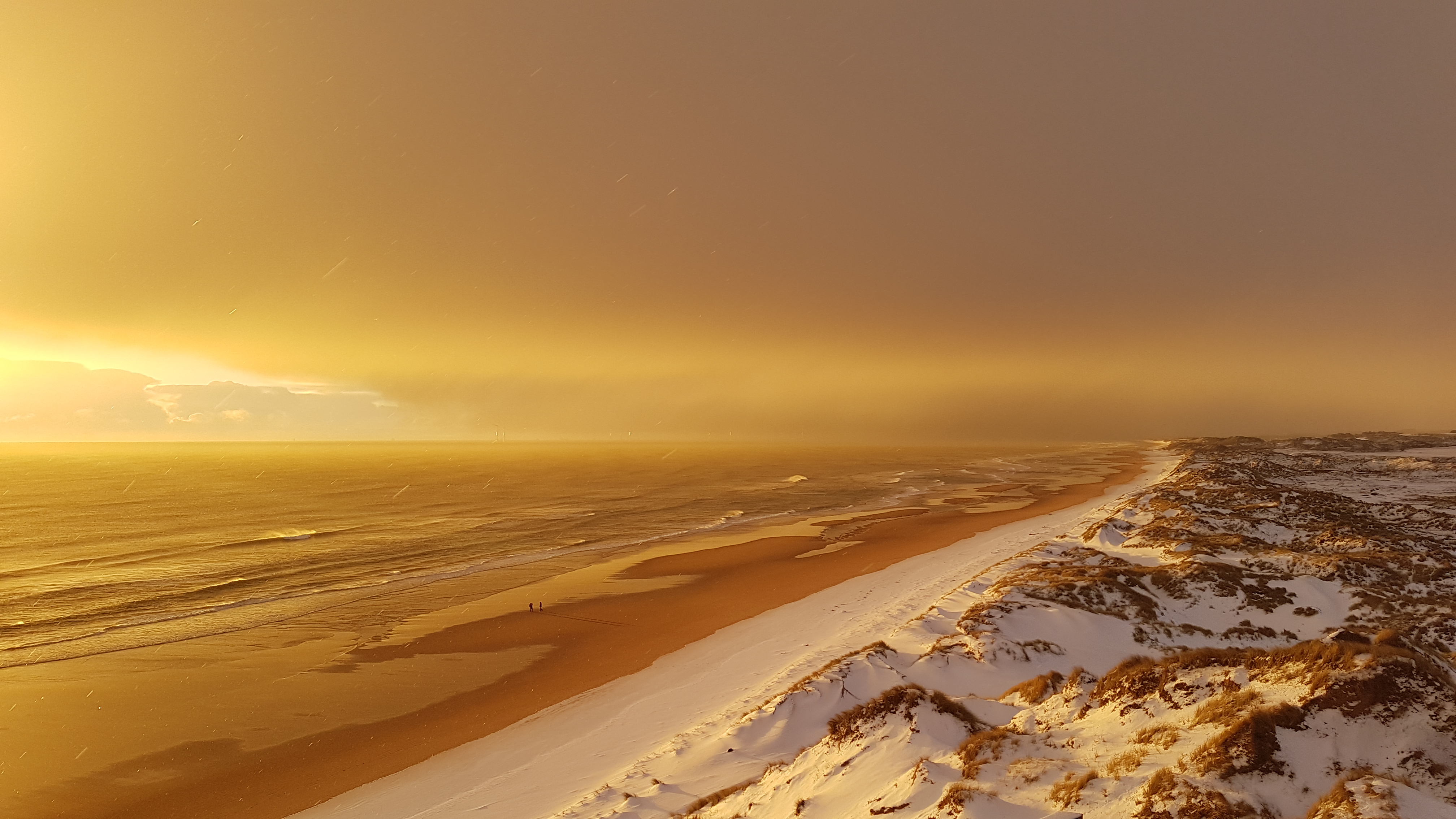A wide golden sunset over a long, empty beach bordered by snow-covered dunes. Soft waves touch the shoreline while warm light breaks through heavy clouds, casting a surreal amber glow across the landscape.