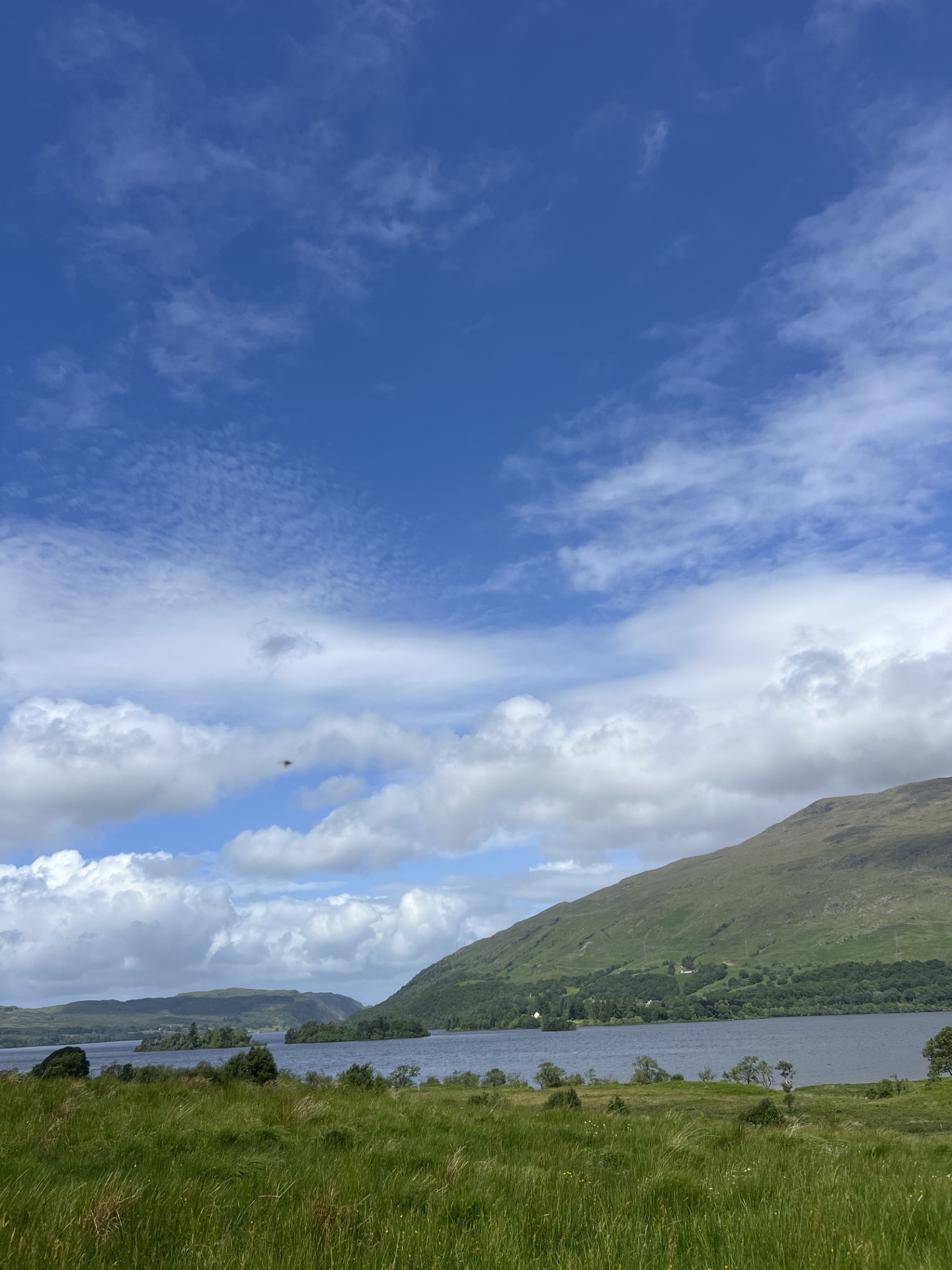 A calm Scottish loch with green hills under a blue, cloud-filled sky.
