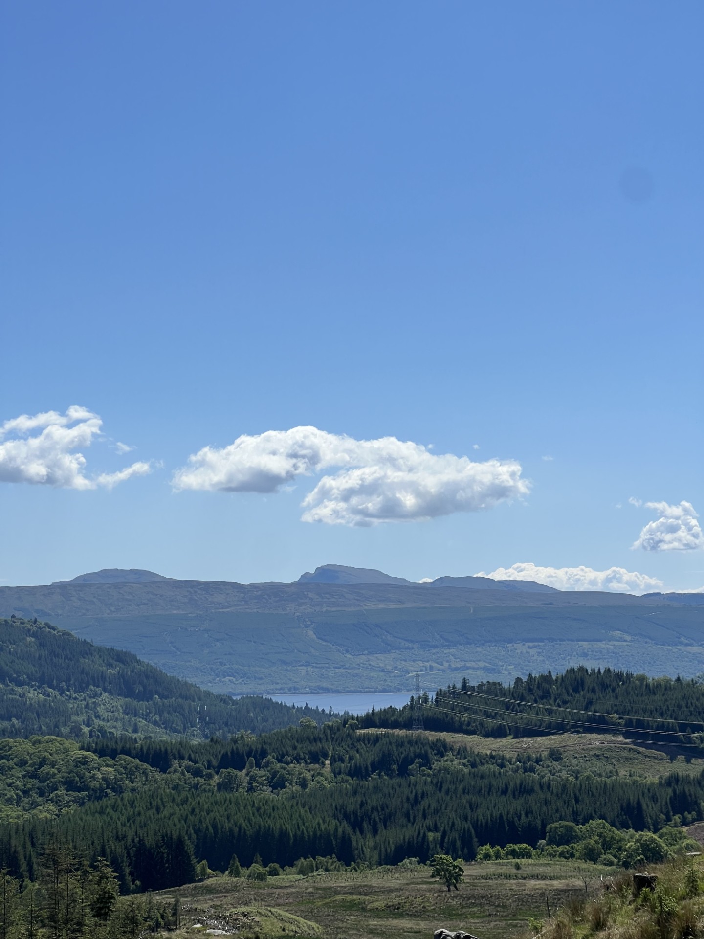 A calm Scottish loch with green hills under a blue, cloud-filled sky.