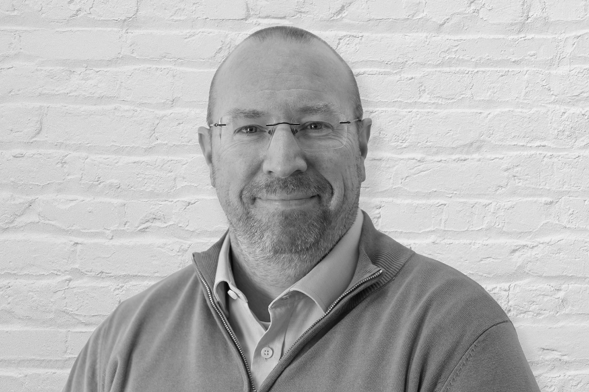 Professional headshot in black and white of a male with blue eyes, shaved brown hair, wearing glasses and a quarter zip fleece with a light blue button shirt underneath