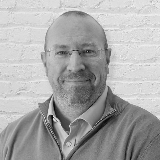 Professional headshot in black and white of a male with blue eyes, shaved brown hair, wearing glasses and a quarter zip fleece with a light blue button shirt underneath