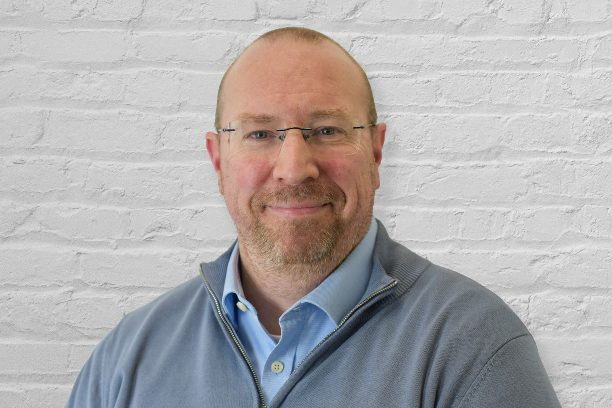Professional headshot in colour of a male with blue eyes, shaved brown hair, wearing glasses and a quarter zip fleece with a light blue button shirt underneath