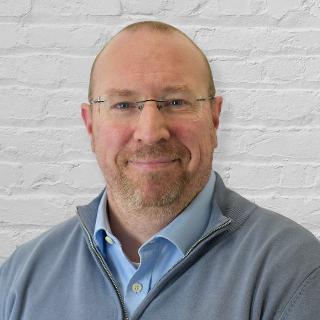Professional headshot in colour of a male with blue eyes, shaved brown hair, wearing glasses and a quarter zip fleece with a light blue button shirt underneath
