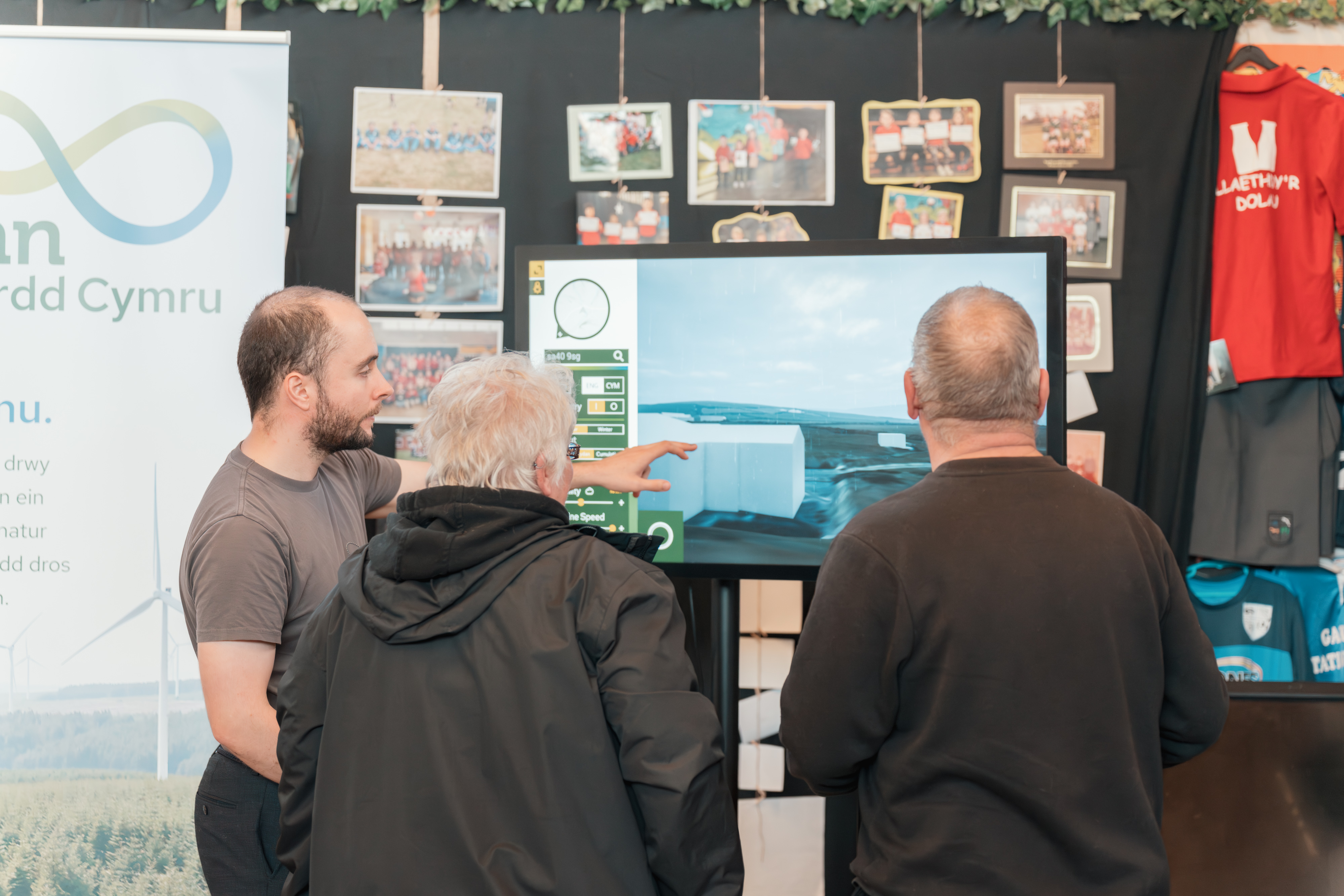 Three people looking at a large screen displaying a digital architectural or landscape model at a community event.