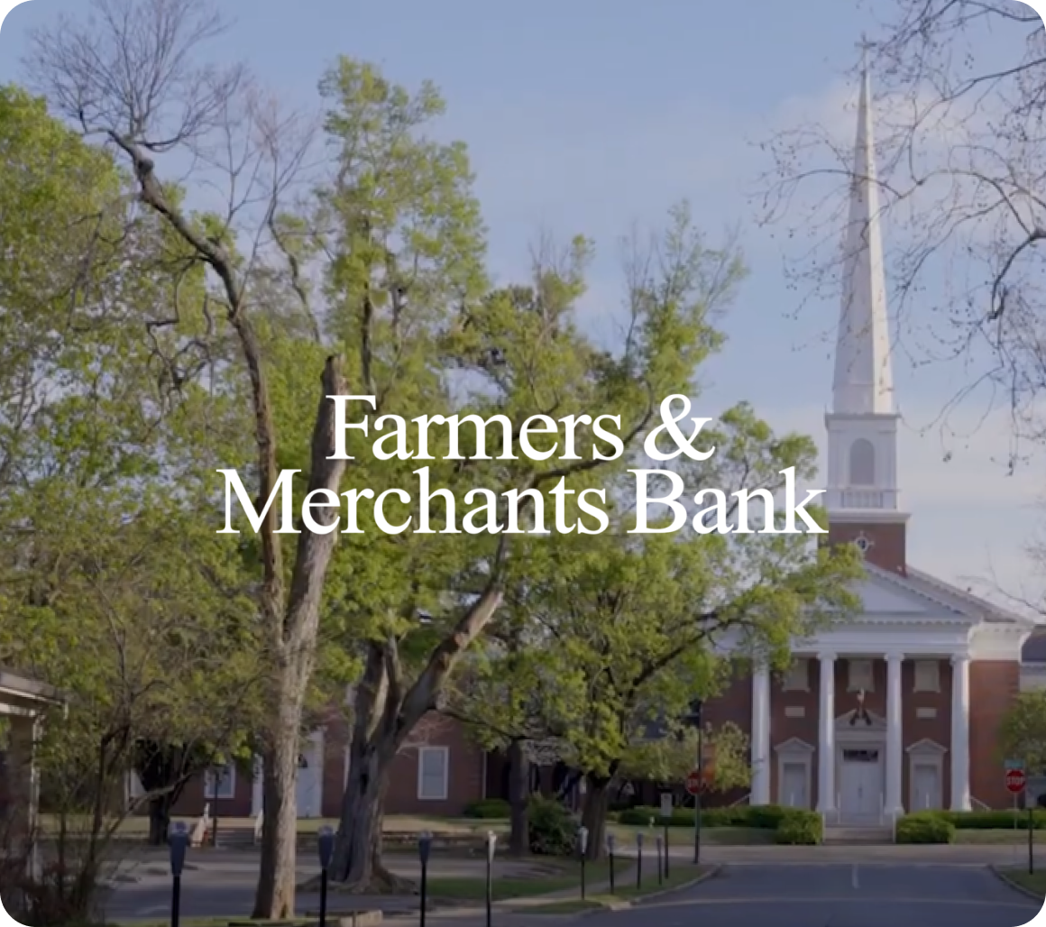 Exterior view of a red-brick building with white columns and a tall steeple surrounded by trees, with the text “Farmers & Merchants Bank” overlaid.