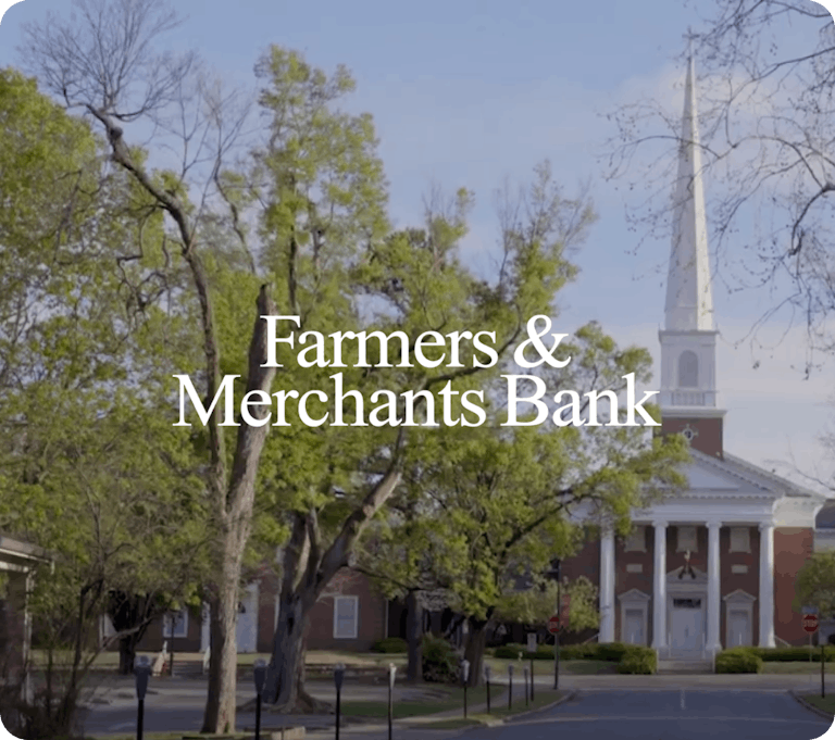 Exterior view of a red-brick building with white columns and a tall steeple surrounded by trees, with the text “Farmers & Merchants Bank” overlaid.