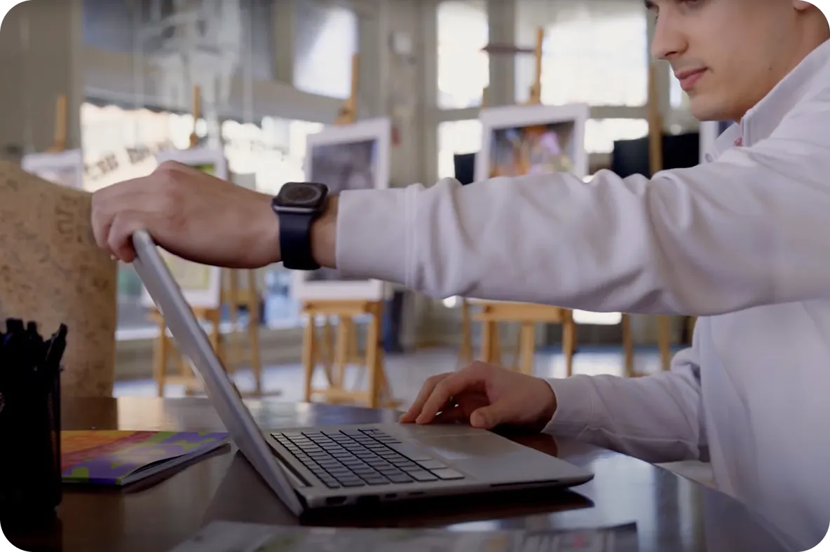 Person closing a laptop while seated at a desk in a bright room with framed artwork in the background.