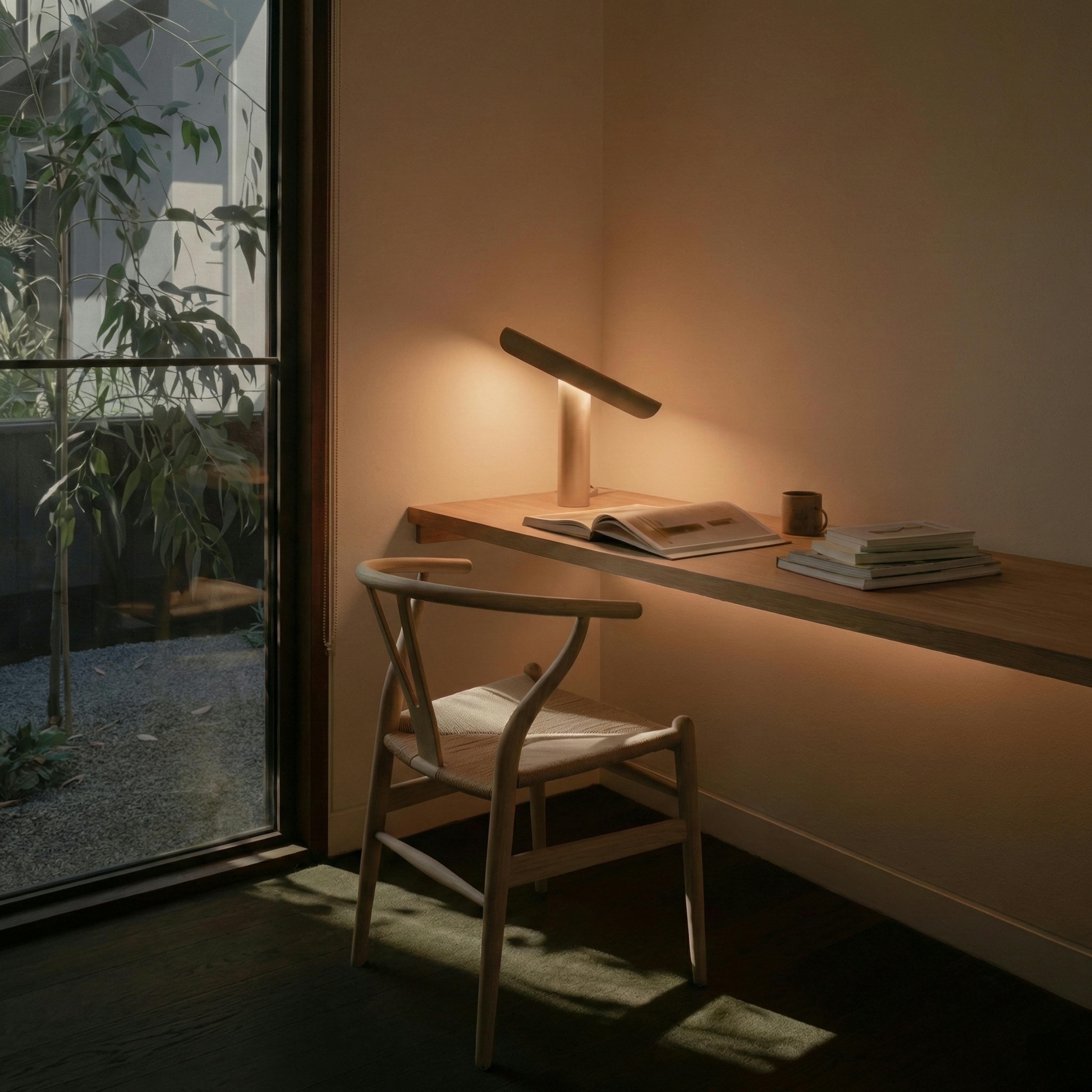 A serene, minimalist home office workspace bathed in intelligent, warm lighting. A light wood floating desk holds an open book under the focused glow of a sleek, modern lamp, paired with a classic woven Wishbone chair. The soft ambient lighting and view of lush garden greenery through the glass door create an atmosphere of simplicity, focus, and holistic wellness achieved through human centric lighting and circadian optimization.