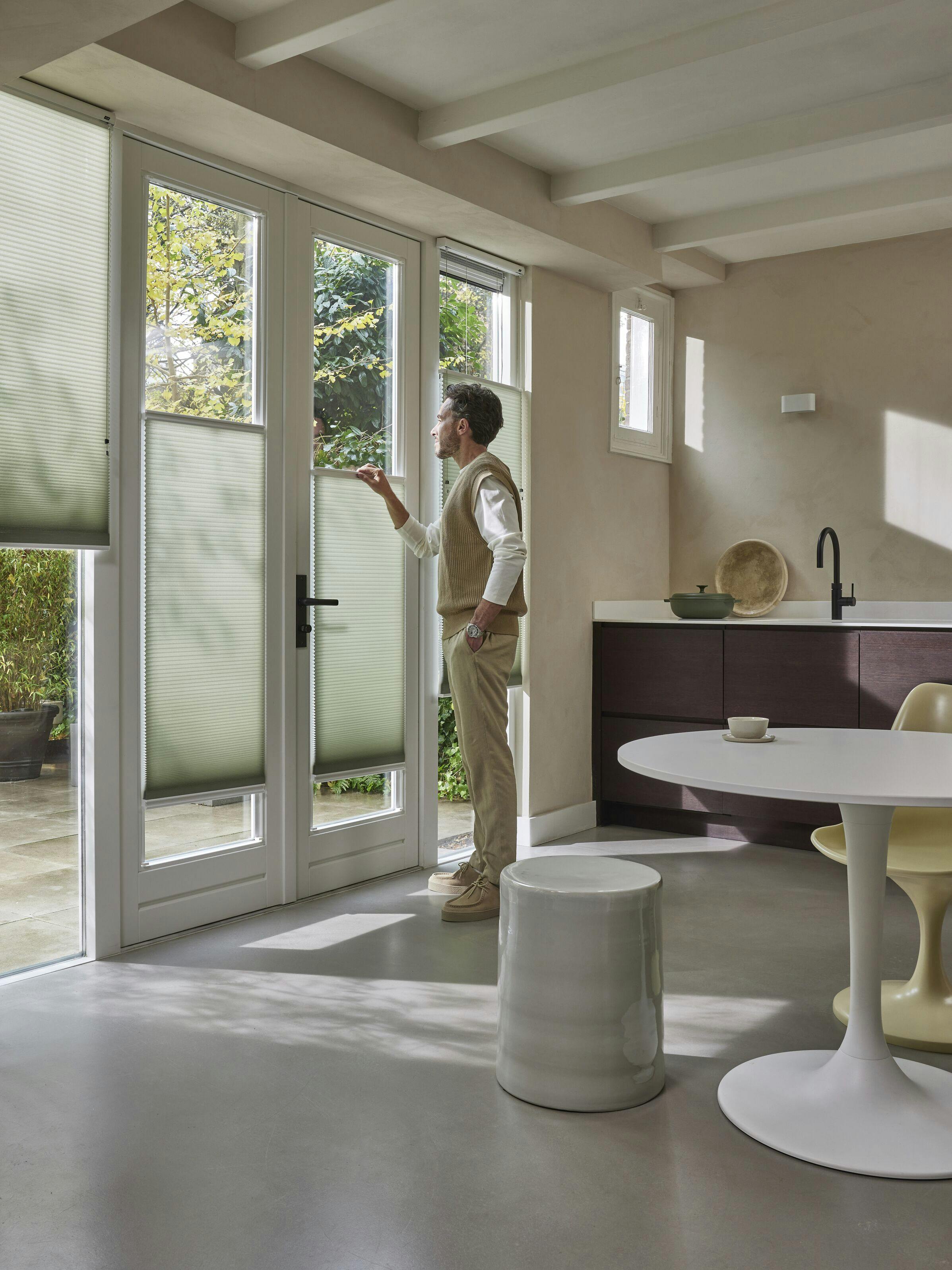 A man standing next to a patio door with top down/bottom up light green Duette® blinds on them in a modern kitchen