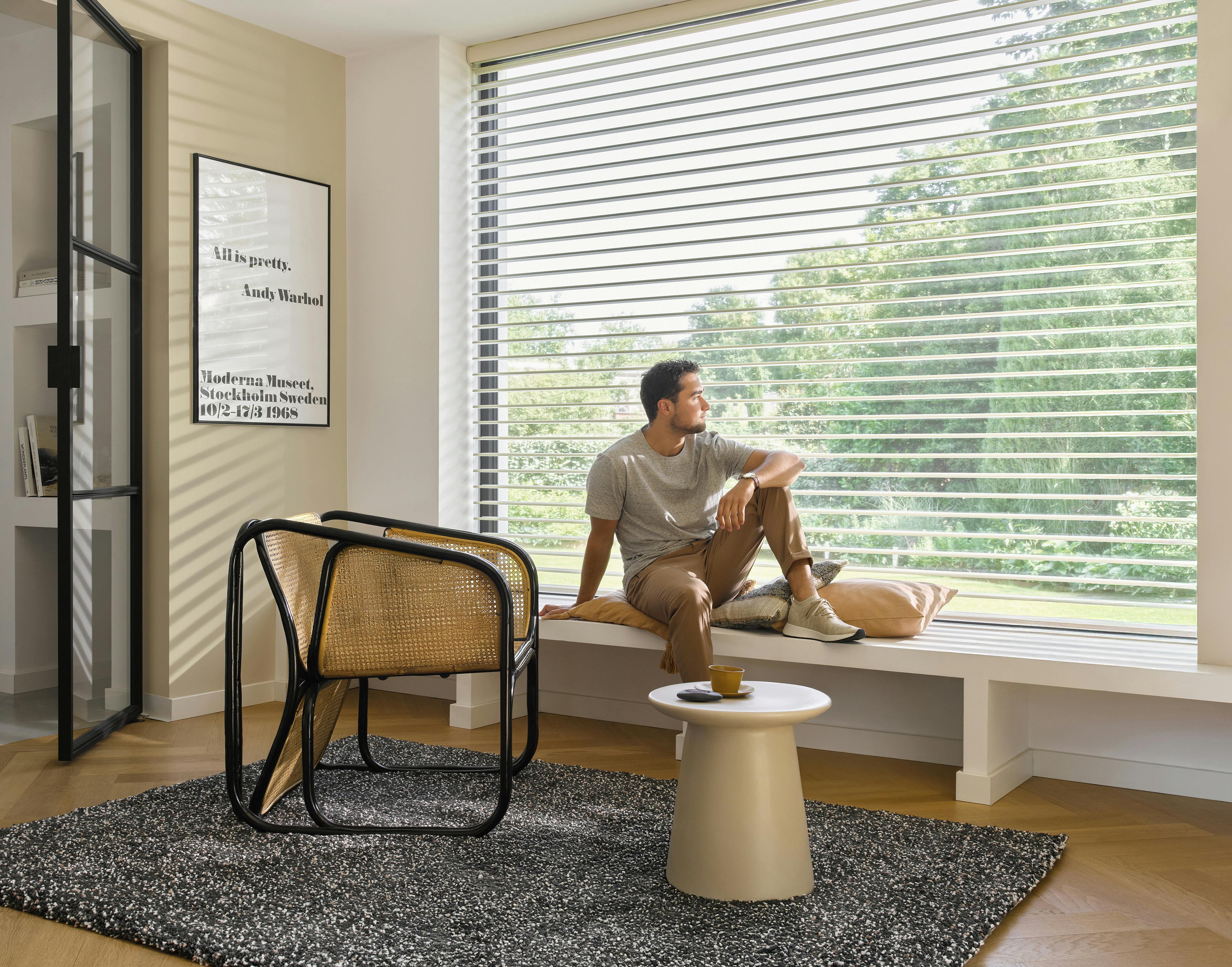 White silhouette shades on a large living room window while a man is sitting by the windowsill