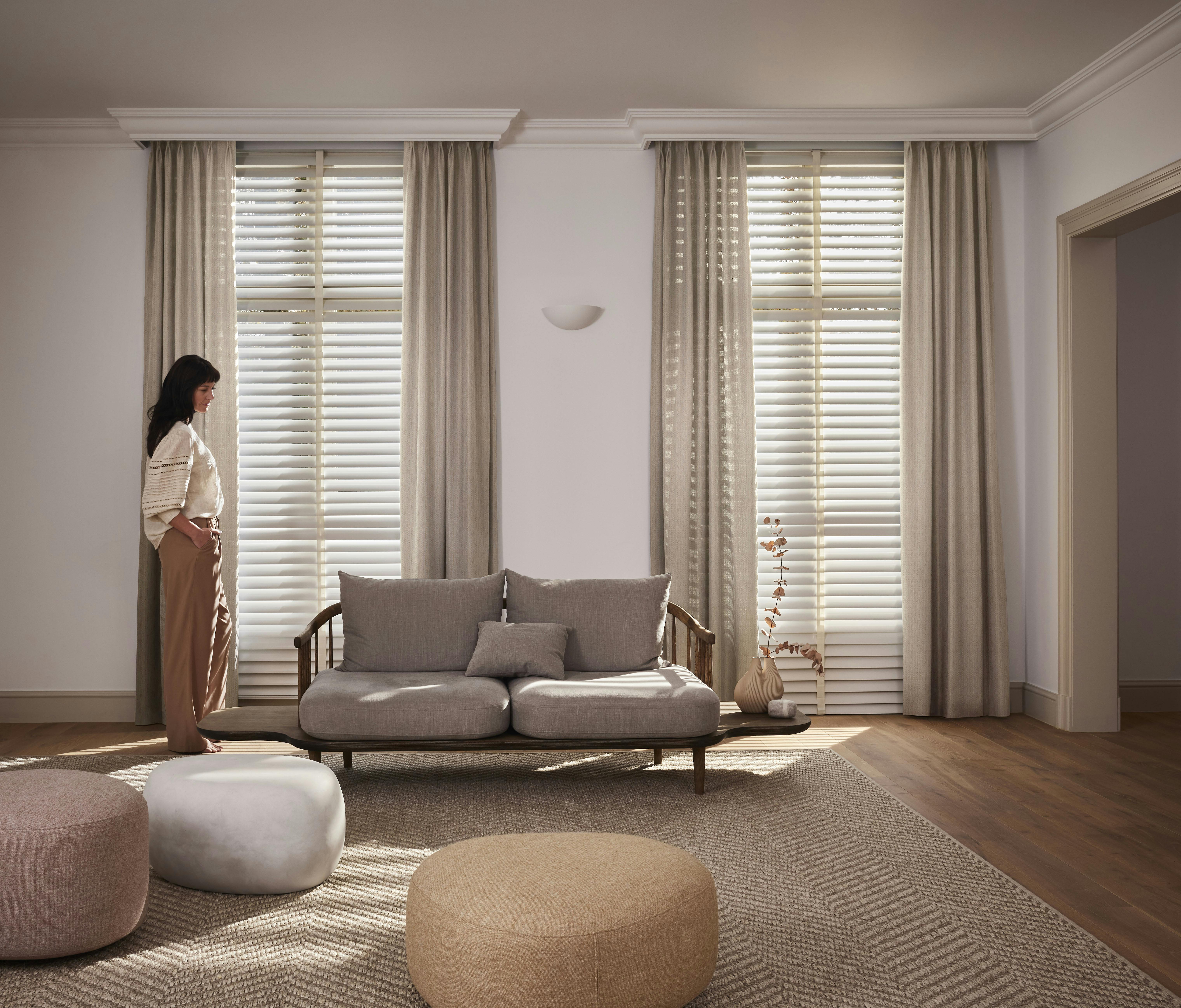 Een vrouw voor ramen met beige gordijnen en witte jaloezieën in een gezellige woonkamer