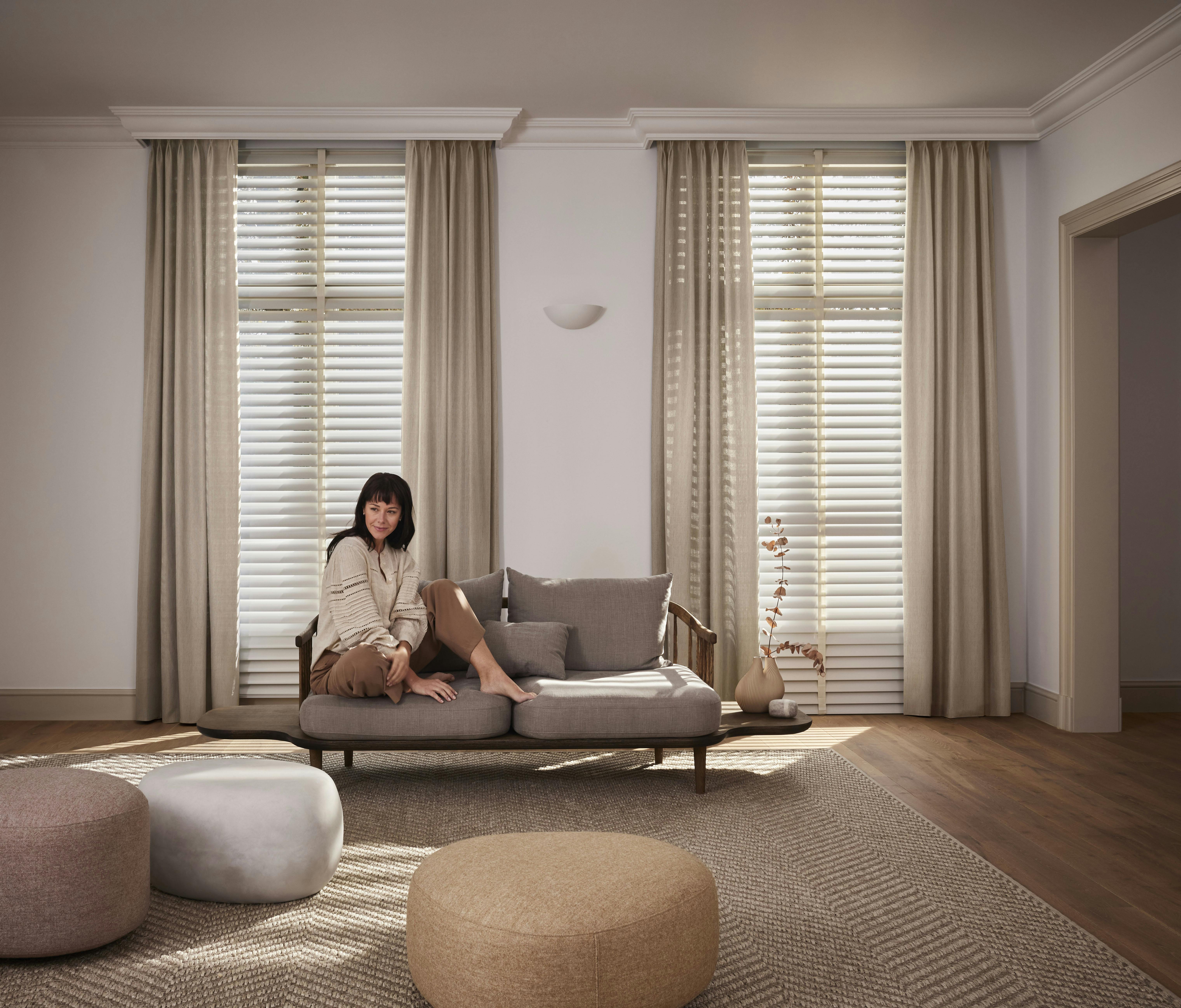 Een vrouw zit op een bank in een gezellige woonkamer met beige gordijnen en witte jaloezieën voor de ramen
