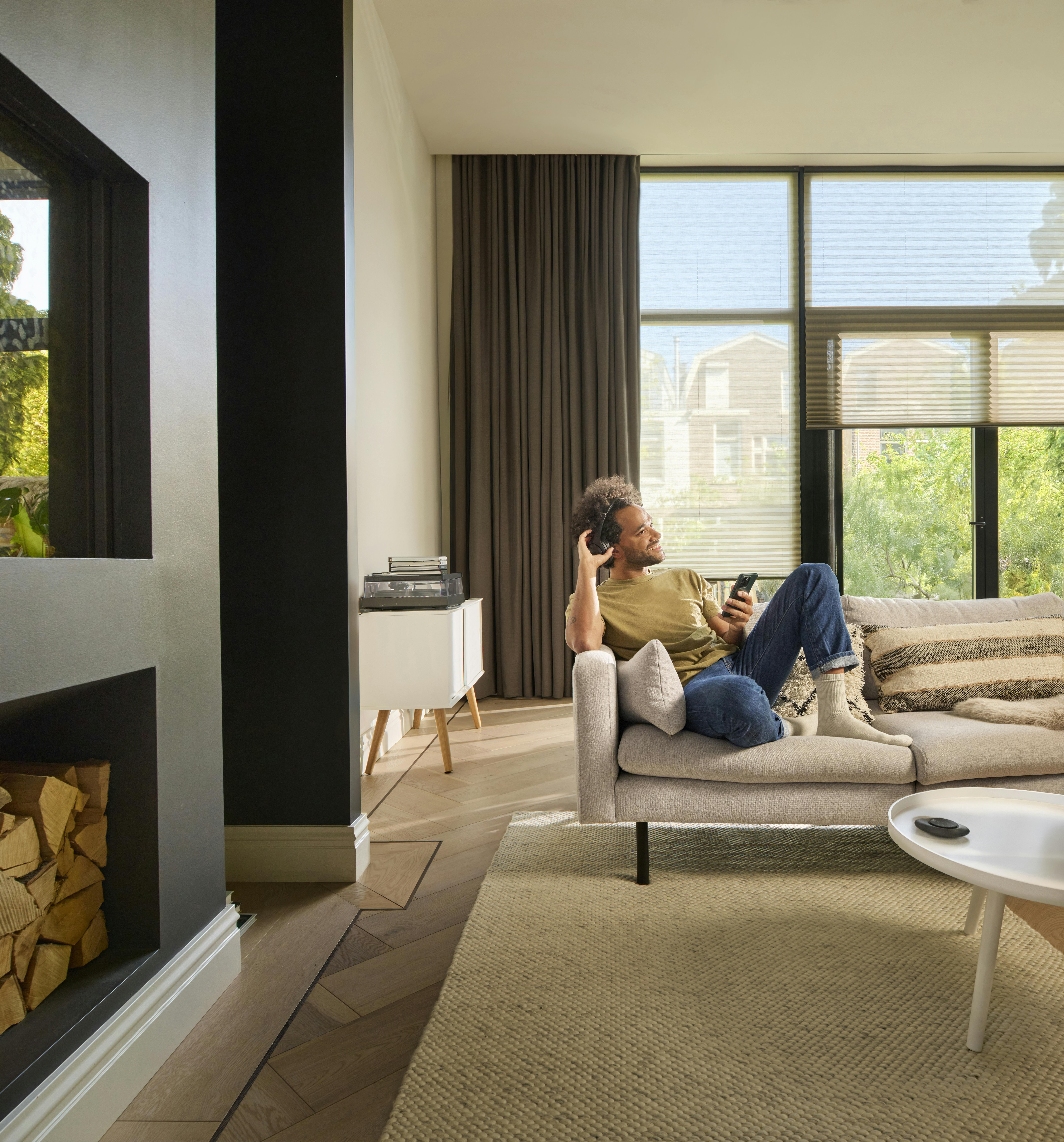 Mand sidder i stuen med brune gardiner og hvide transparente plisségardiner, styret med Pebble® fjernbetjening til PowerView® automation