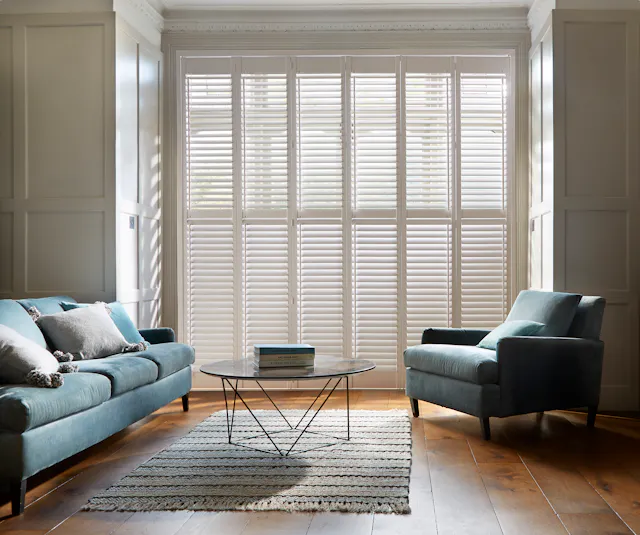 Witte shutters op grote ramen in een moderne woonkamer met een blauwe zetel en armstoel