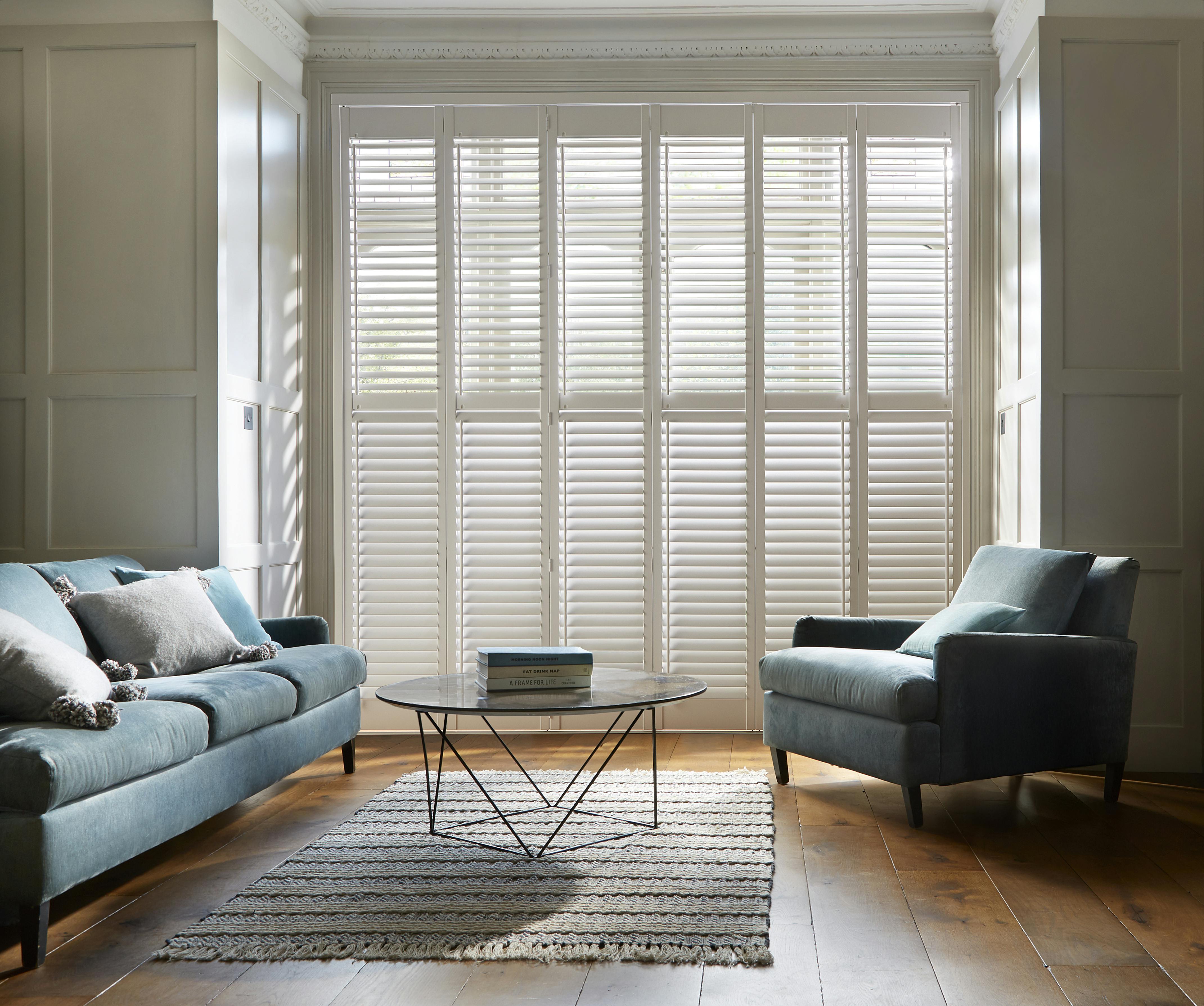White shutters on large windows in a modern living room with blue sofa and an armchair
