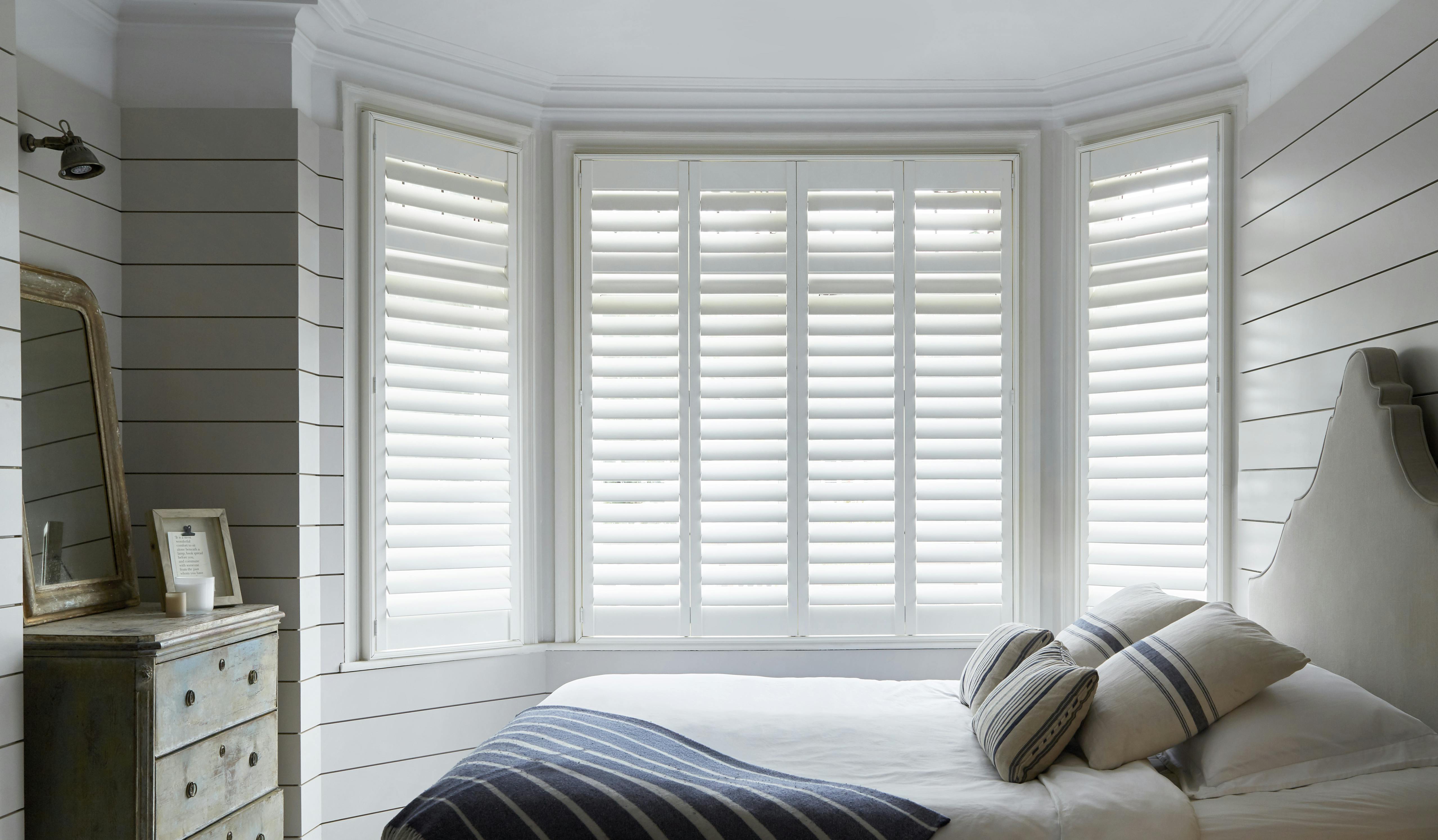 White shutters on a bay window in a light cosy bedroom 