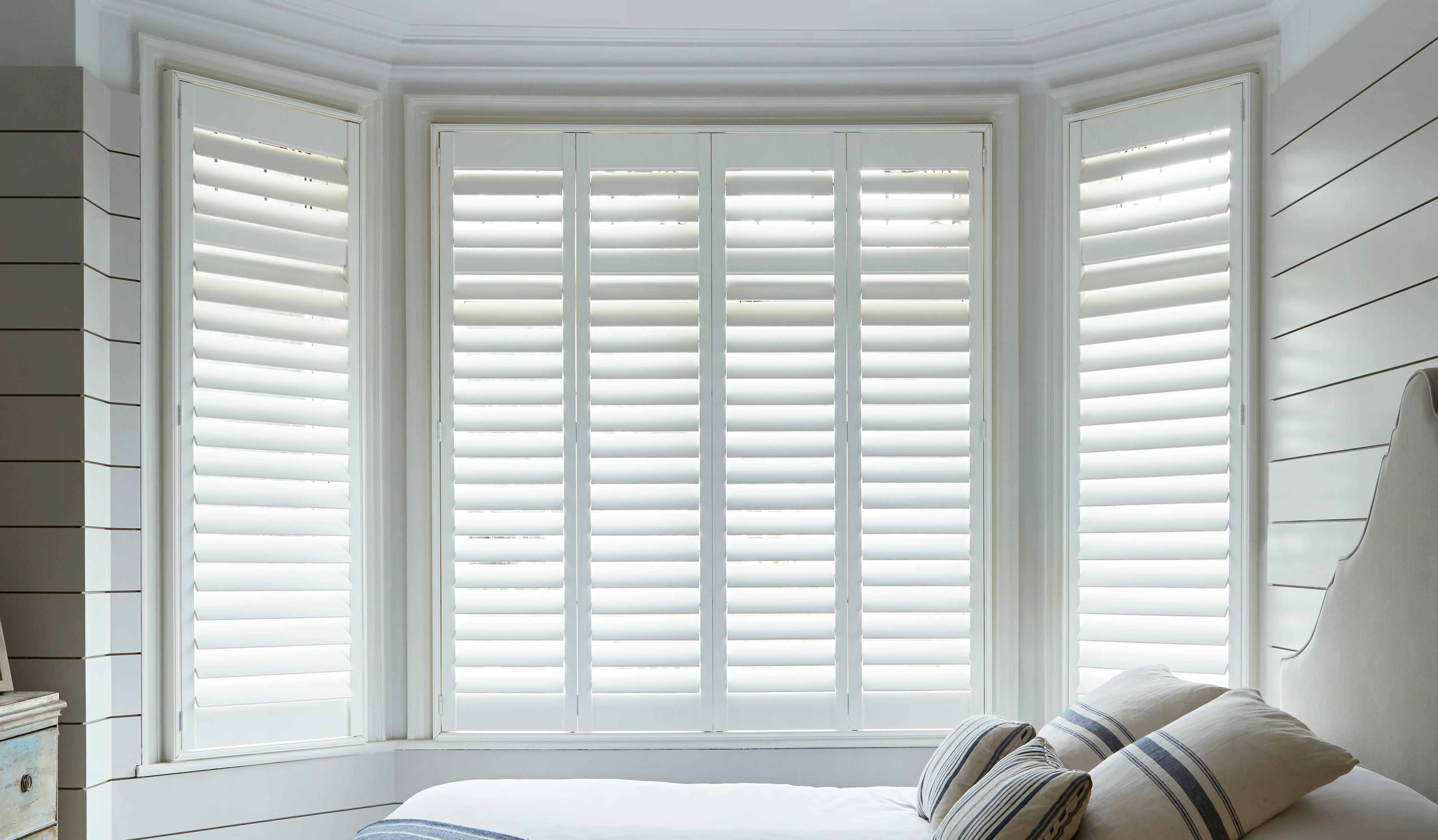White shutters on a bay window in a light cosy bedroom
